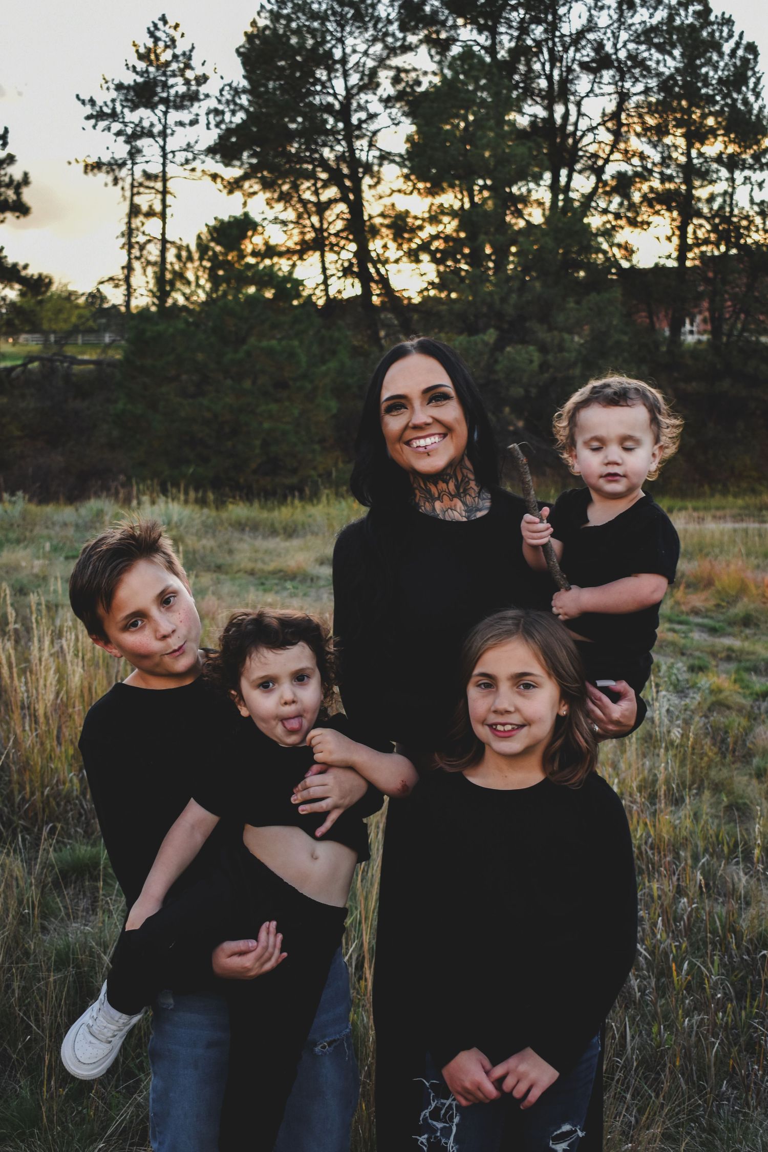 A laughing mother in black holds a toddler while her three children make playful faces in a sunset field.