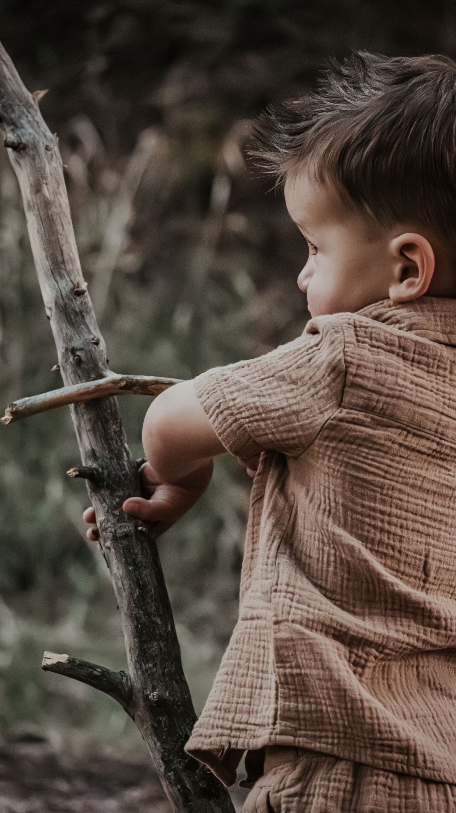 Young toddler boy in a beige textured shirt holding a wooden stick branch outdoors in nature.