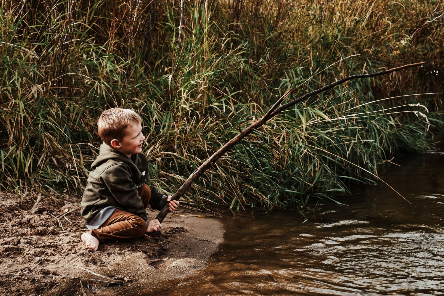 Young boy sits by muddy riverbank holding long stick while fishing in tall grass surroundings. Family Photographer in Pflugerville, Tx