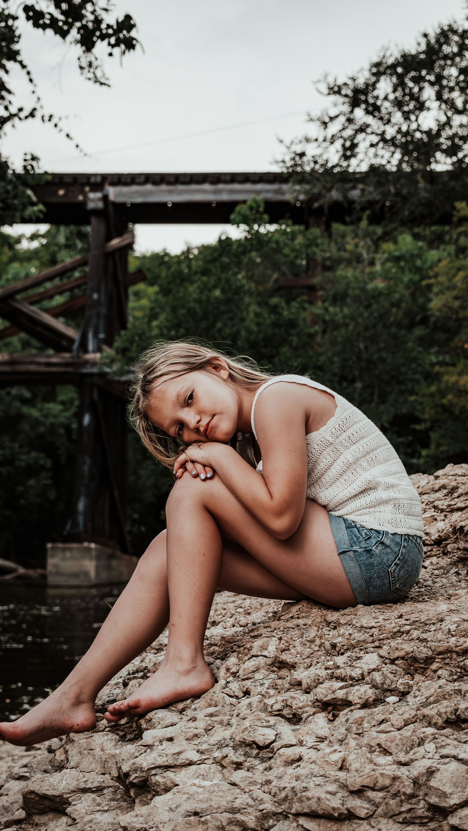 Barefoot blonde girl sitting on rocky surface near a bridge in a wooded outdoor setting.