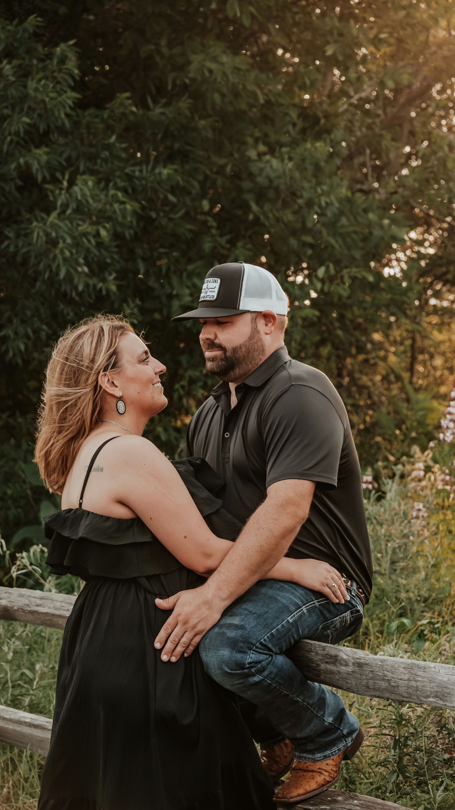 Romantic couple gazing at each other by a wooden fence surrounded by wildflowers at sunset.