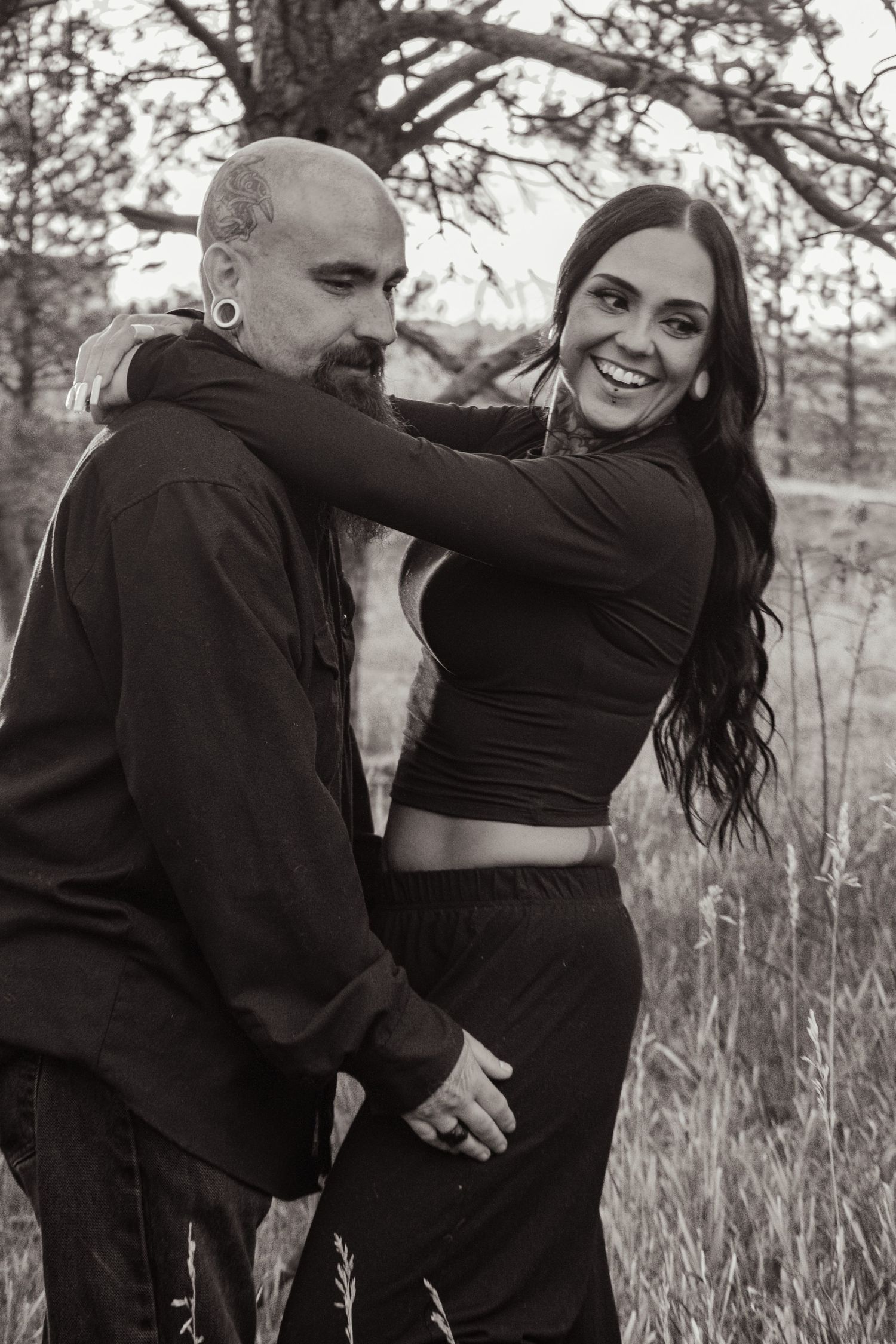 Black and white photo of a smiling couple embracing in a field, woman with long dark hair hugging bald man.