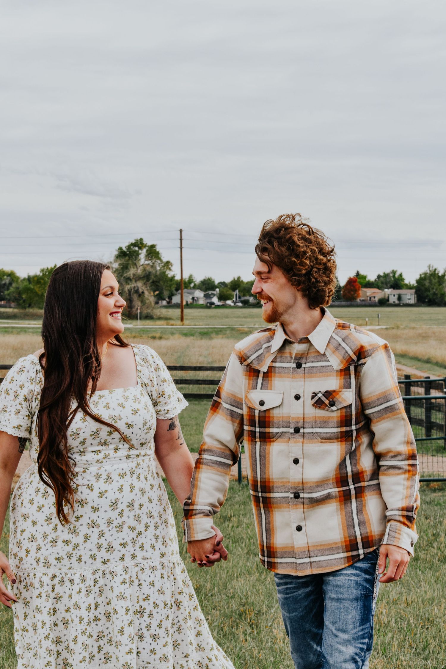 A couple walks hand in hand through a green field, smiling at each other on a cloudy day.