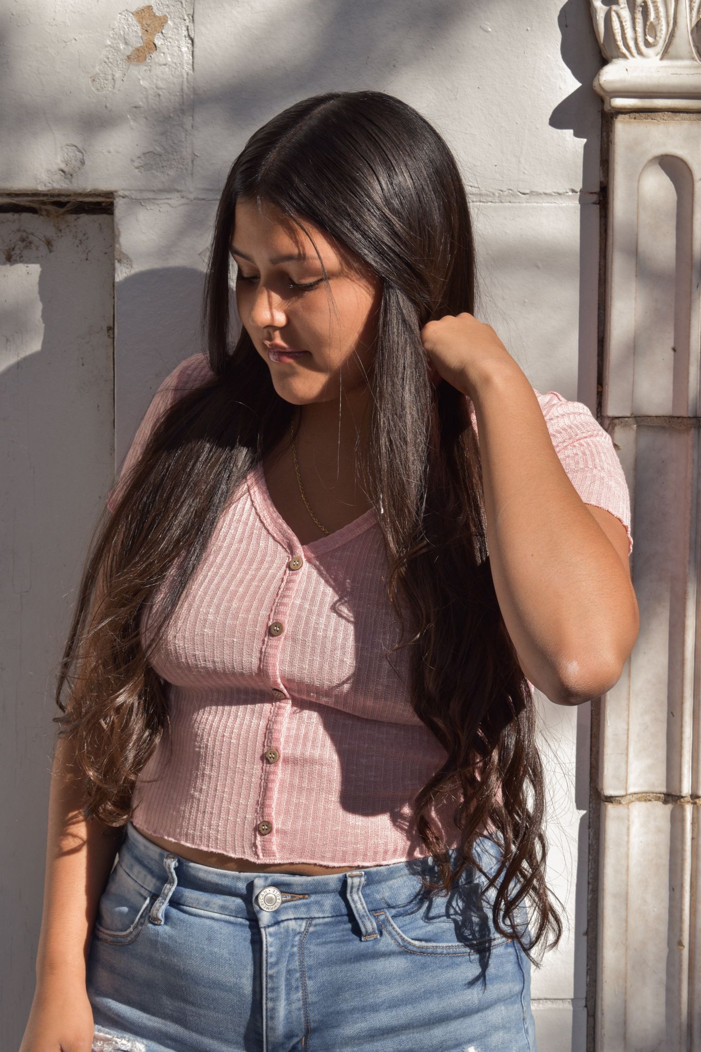 Young woman with long dark hair touches her hair while standing by white wall for senior photos.