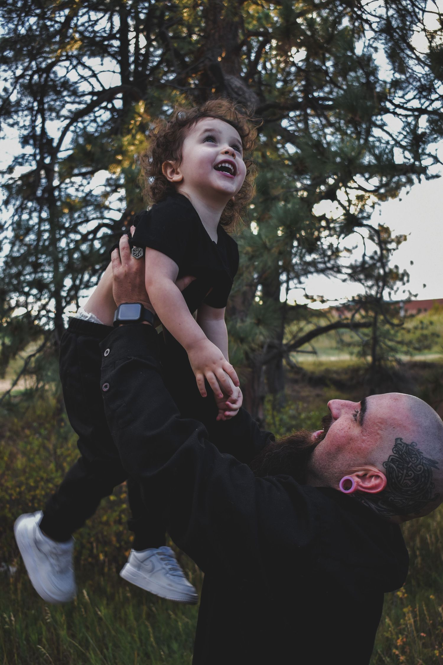 Joyful father lifting laughing curly-haired toddler in the air among pine trees outdoors.