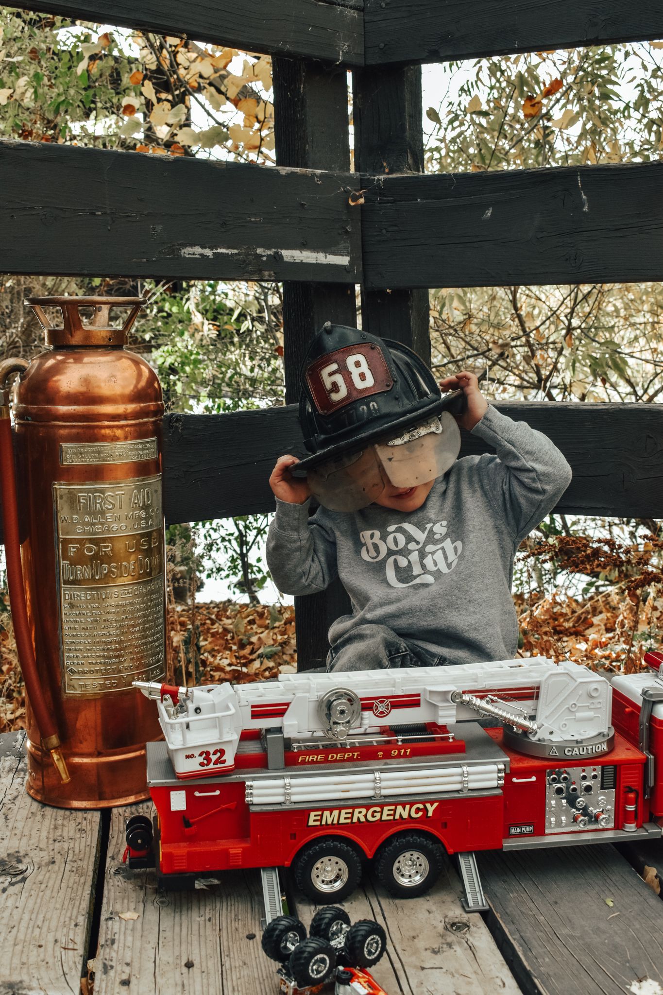 Little boy adjusting black firefighter helmet number 58 beside vintage copper fire extinguisher and toy emergency truck.