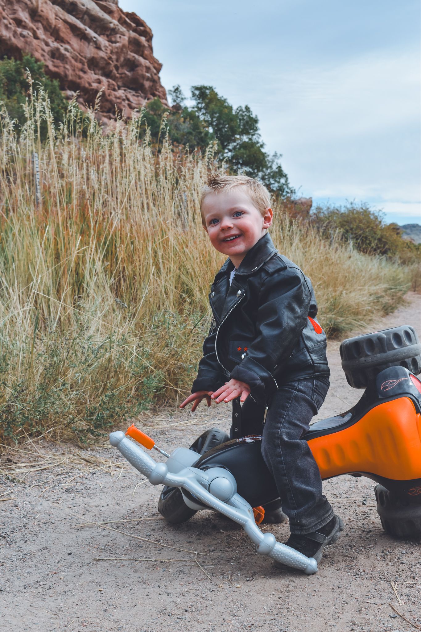 Smiling toddler in black leather jacket riding orange toy motorcycle on dirt path with tall grass.