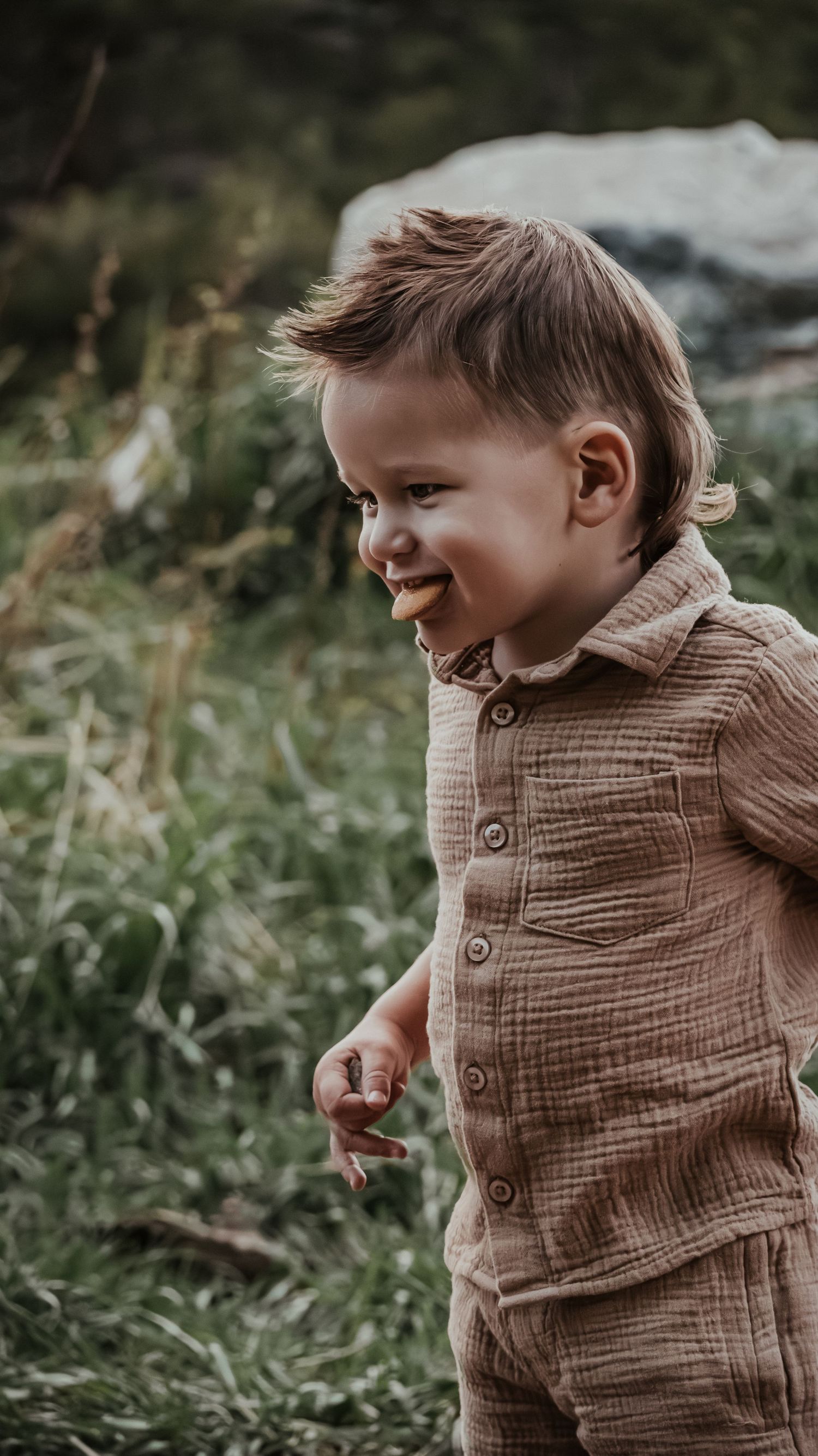 Happy toddler boy in brown linen outfit smiling while playing outdoors in green nature setting.