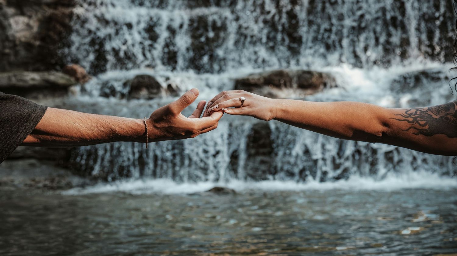 Two tattooed hands gently touching in front of a cascading waterfall.