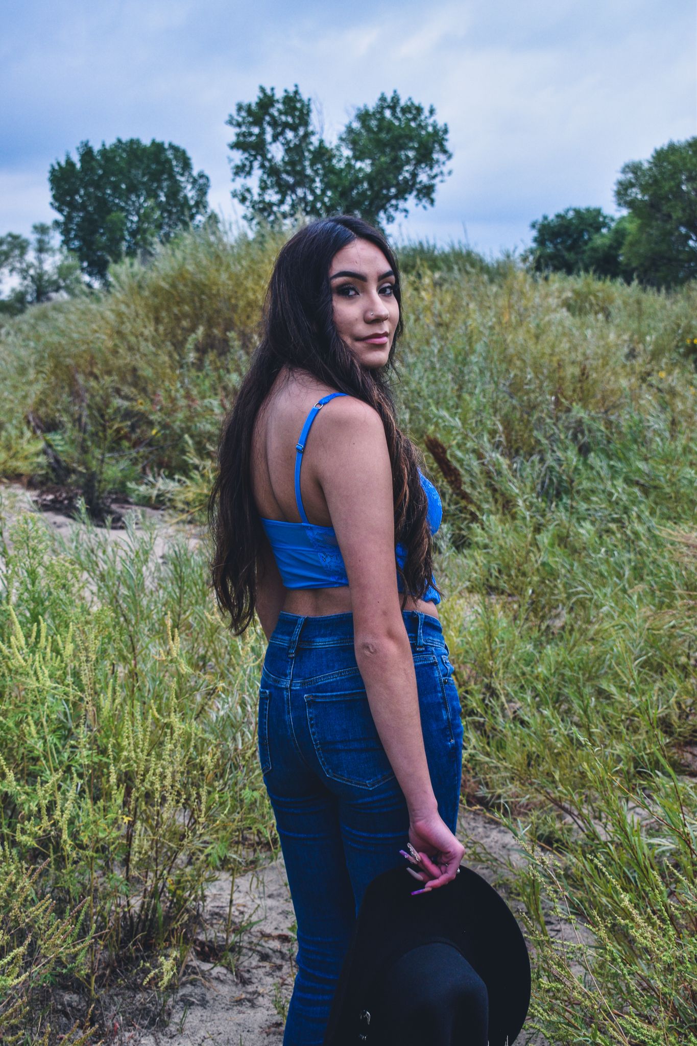 Young woman in blue crop top and jeans holds black hat while posing in a sandy field with greenery for senior photos.