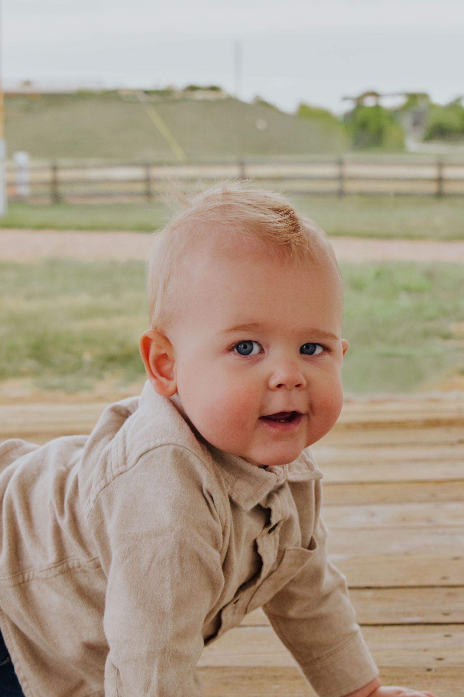 Smiling baby with blue eyes in beige shirt crawling on wooden deck outdoors.