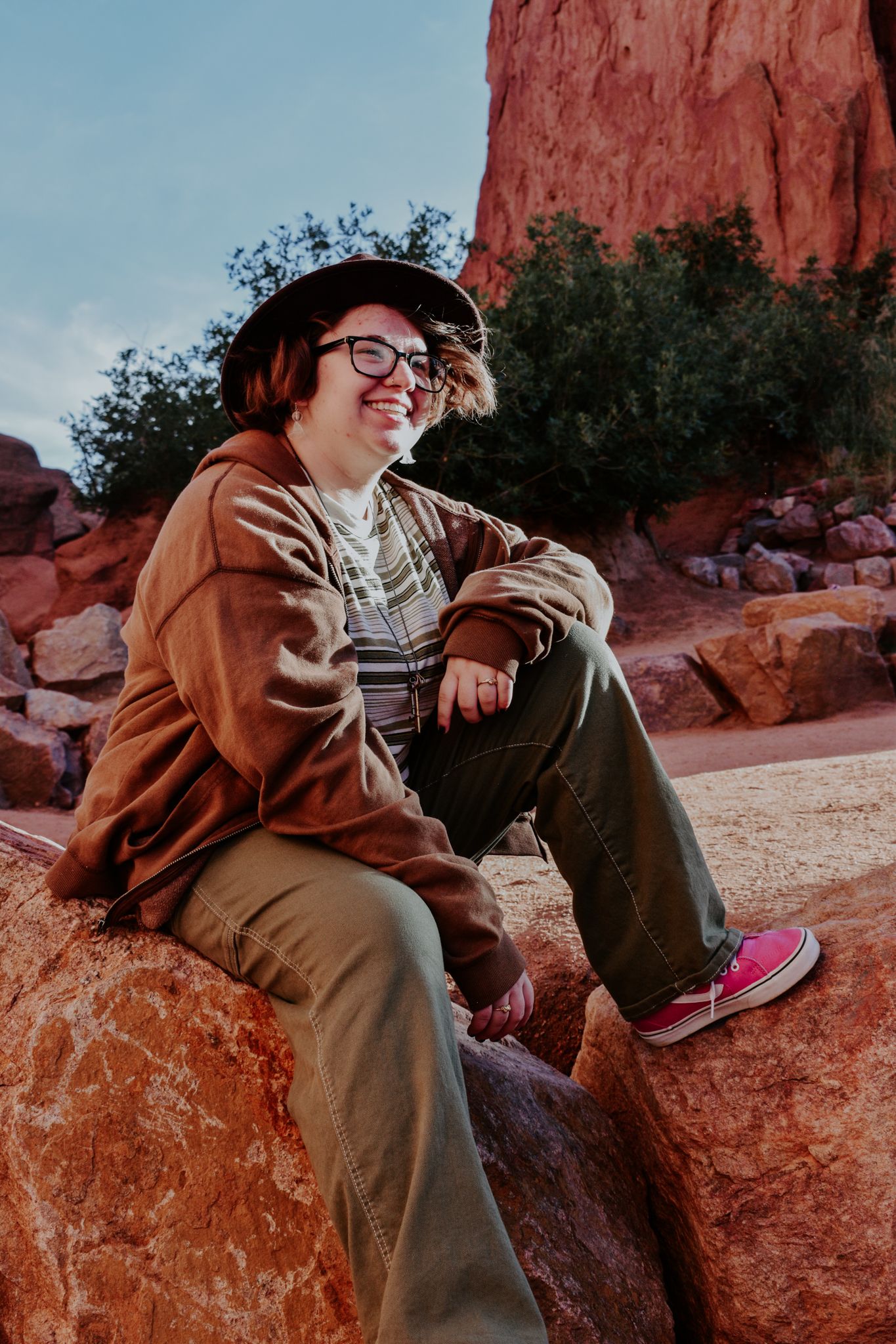 Smiling person in brown jacket, hat, and glasses sitting on red rocks at Garden of the Gods for senior photos.
