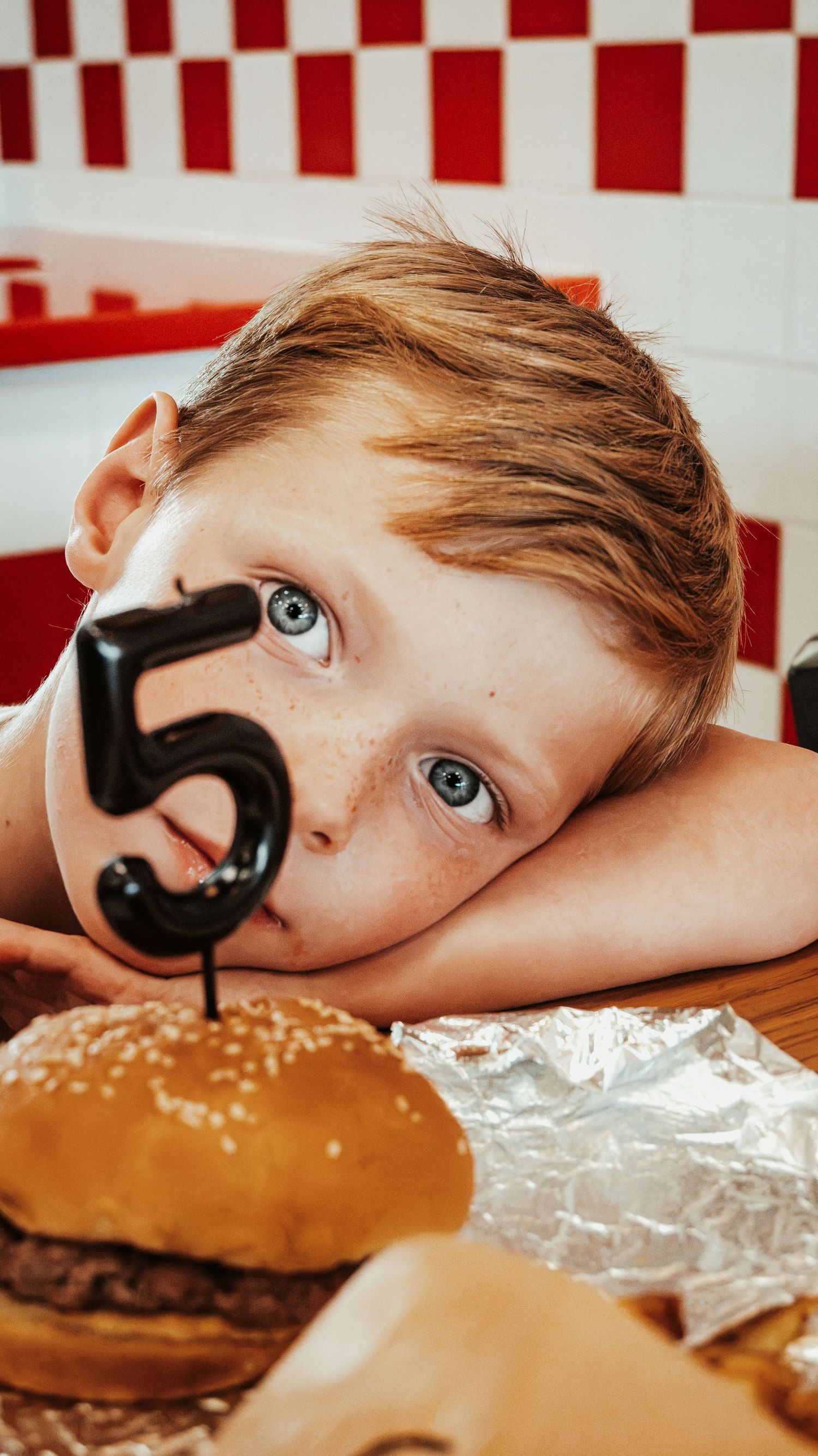 A boy rests his chin on a table next to a burger with a number 5 candle at Five Guys. Family Photographer in Pflugerville, Tx
