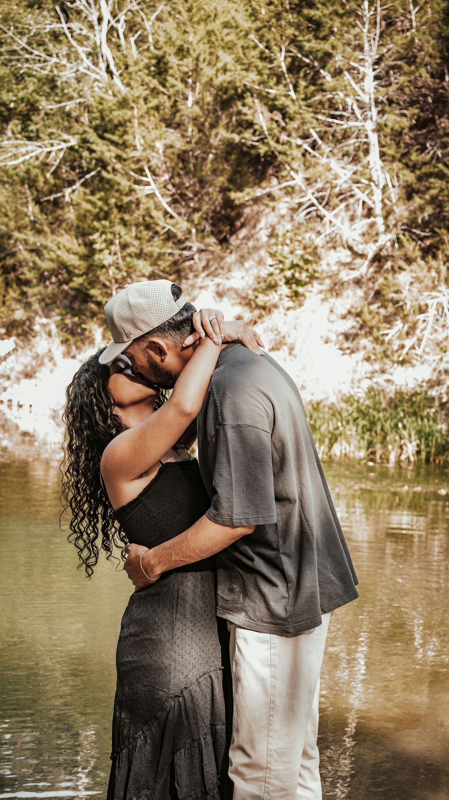 Couple sharing a romantic kiss near a serene lake surrounded by lush green trees for a couples session.