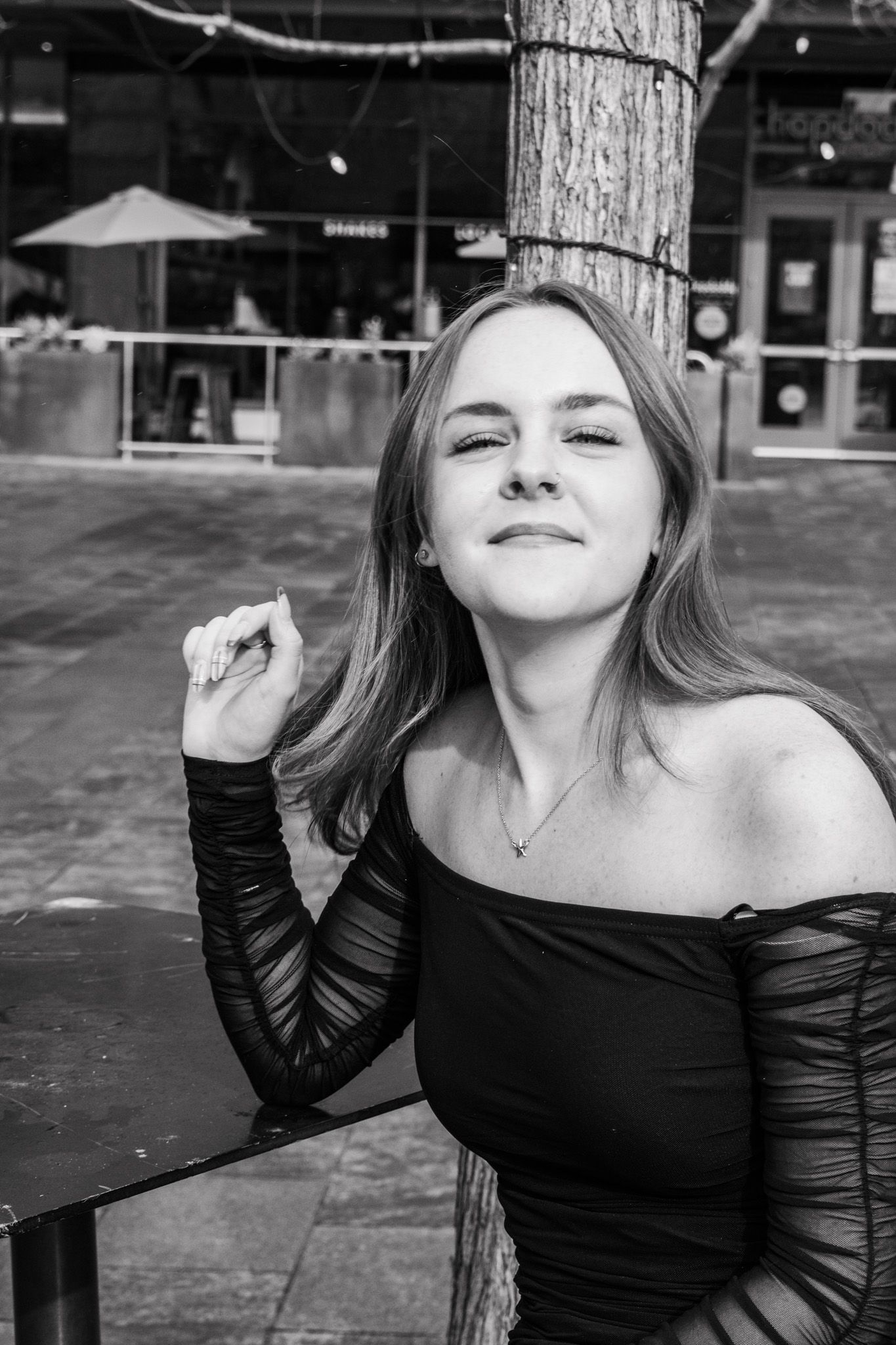 Young blonde woman in black off-shoulder top smiling while leaning against tree in black and white photo for senior photos.