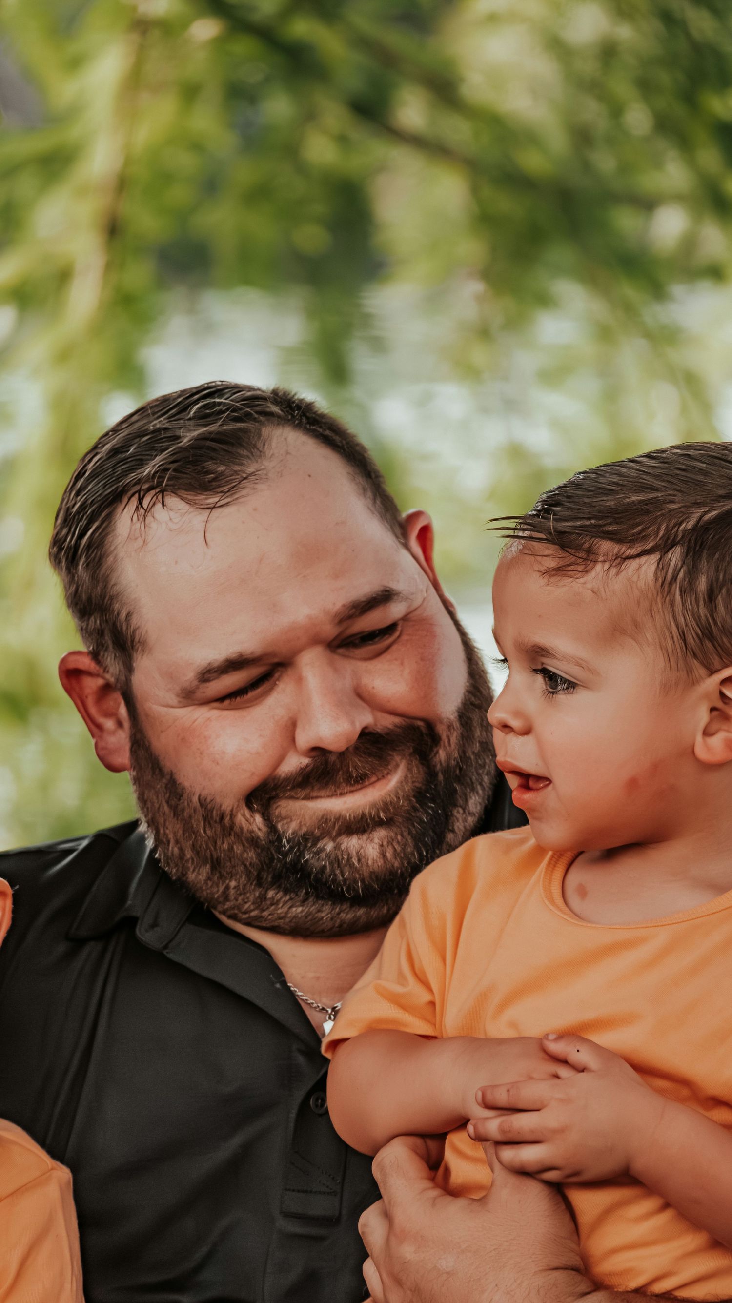 Smiling bearded dad holds his young son in an orange shirt during an outdoor photo session.