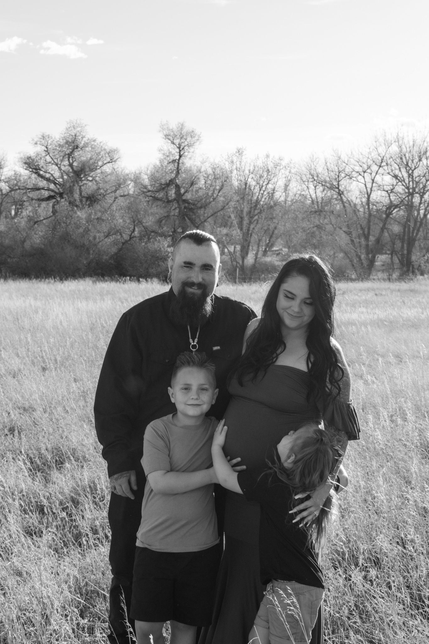 Black and white family maternity photo with father, child, and pregnant mother embracing in a dry grass field.