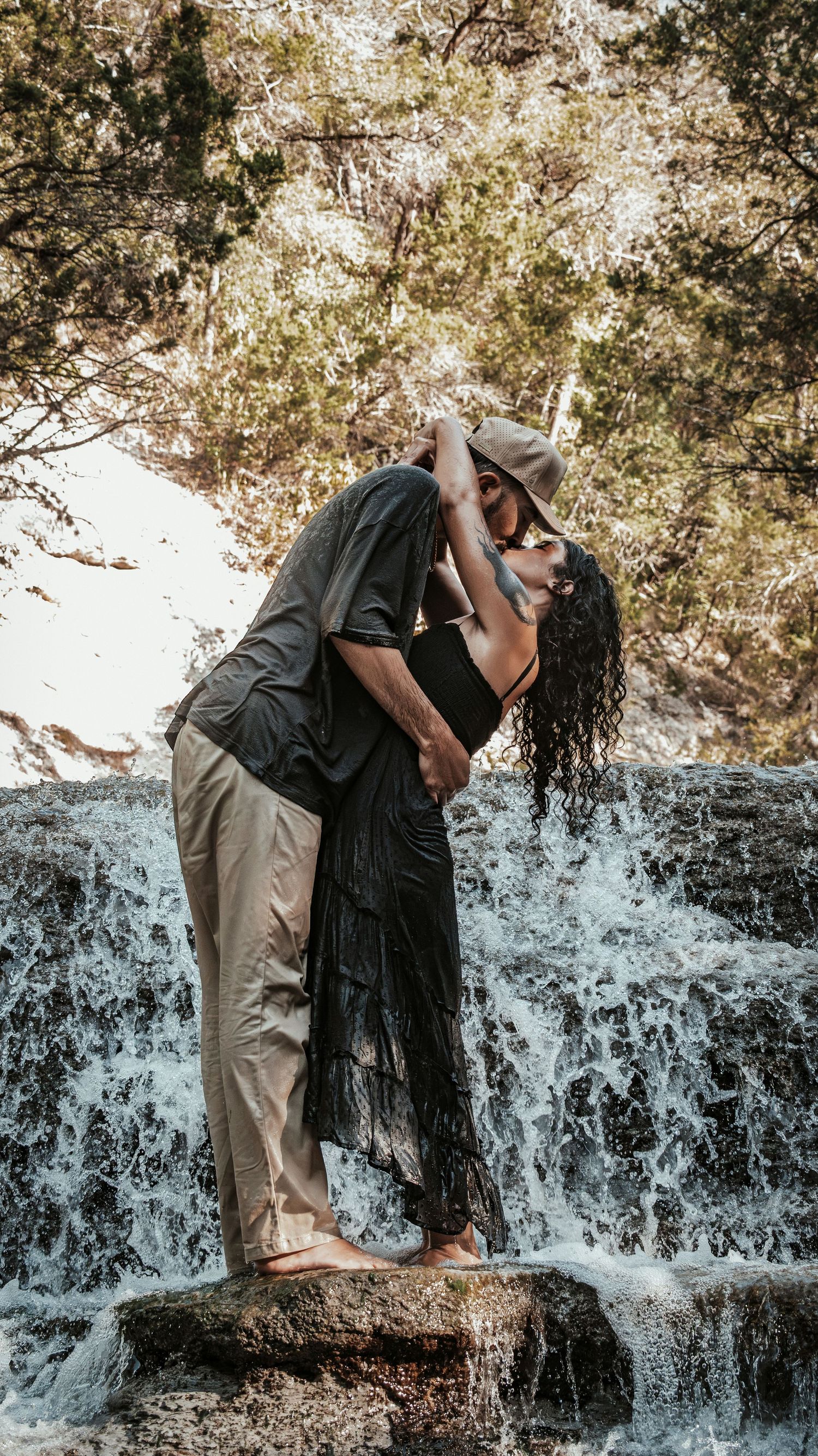 Couple in a romantic dip pose kissing in front of a waterfall, surrounded by lush green trees.