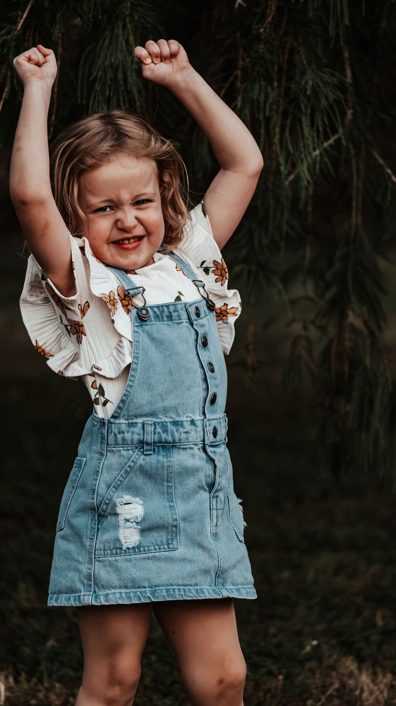 Energetic toddler girl in denim overalls raising fists triumphantly with a fierce expression outdoors.