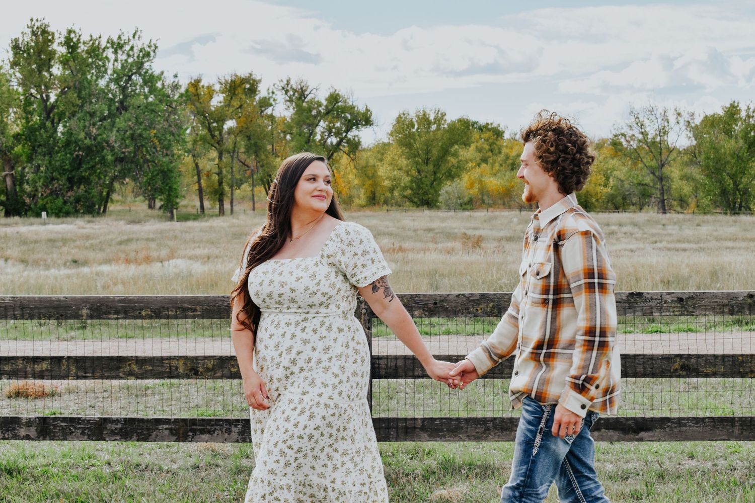 Couple holding hands and gazing at each other near a wooden fence in an autumn farm setting.