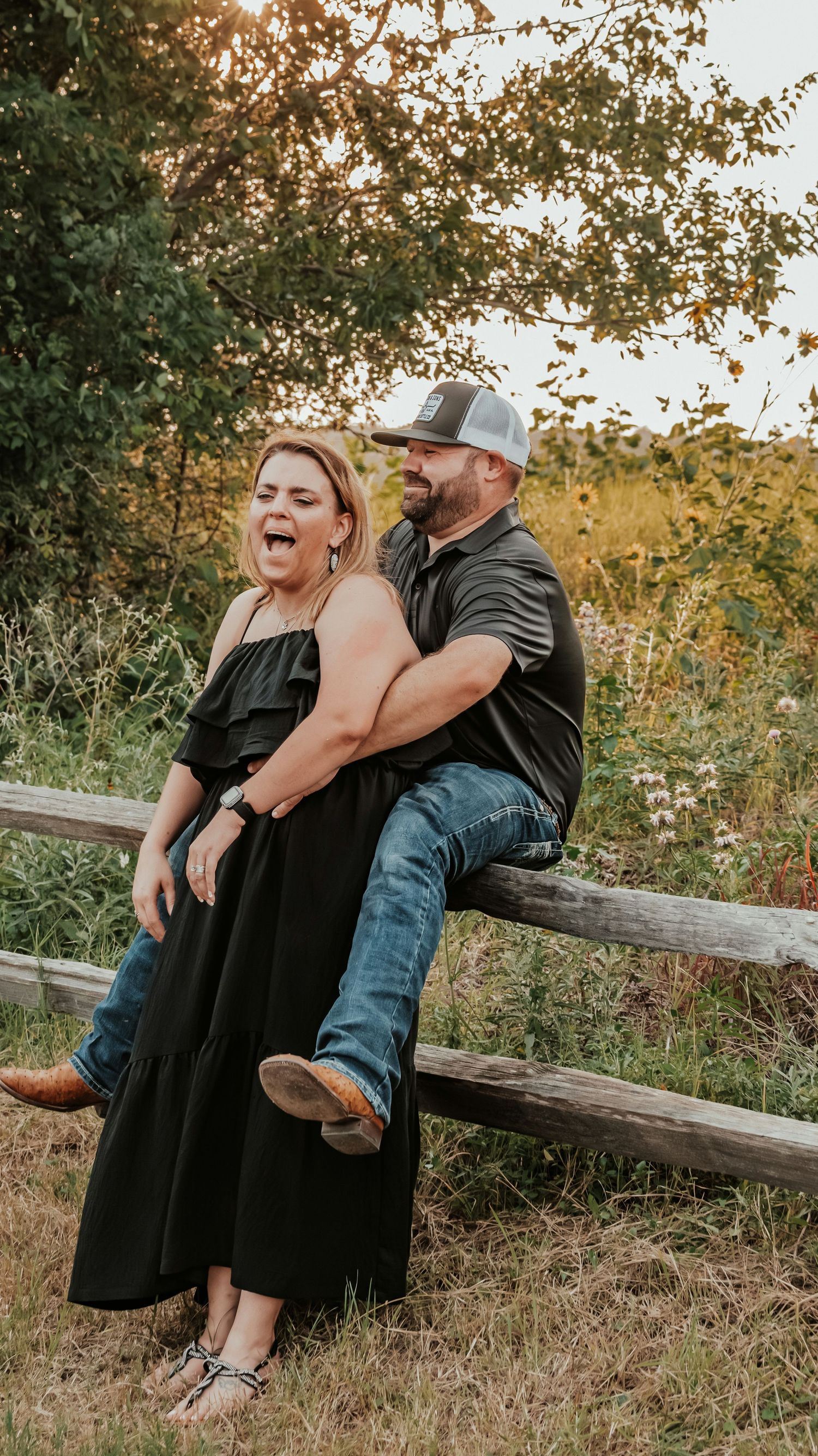 Laughing couple relaxing on a rustic wooden fence in a wildflower meadow at sunset.