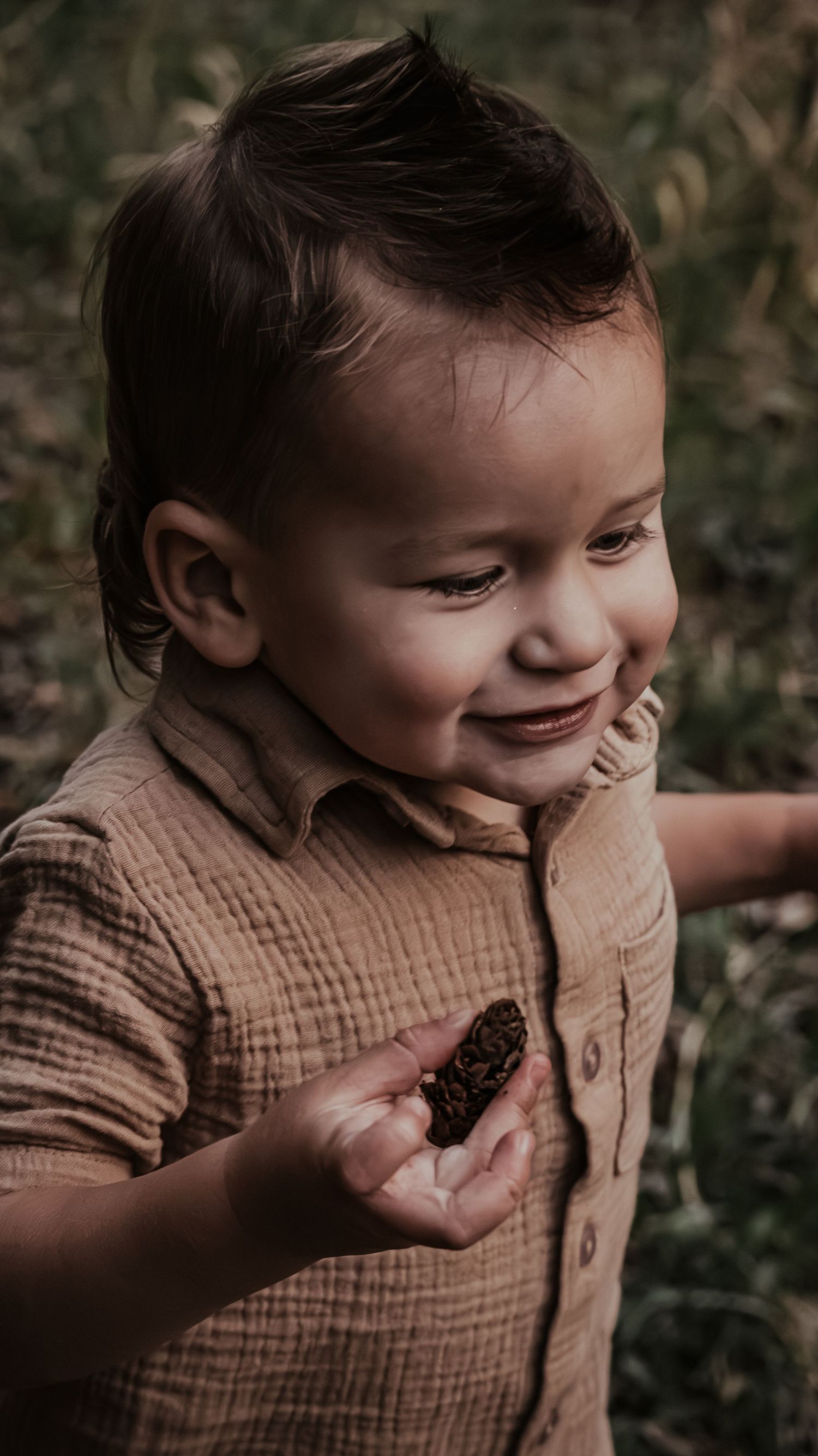 Smiling toddler boy in tan button-up shirt holds a pinecone in his outstretched hand.