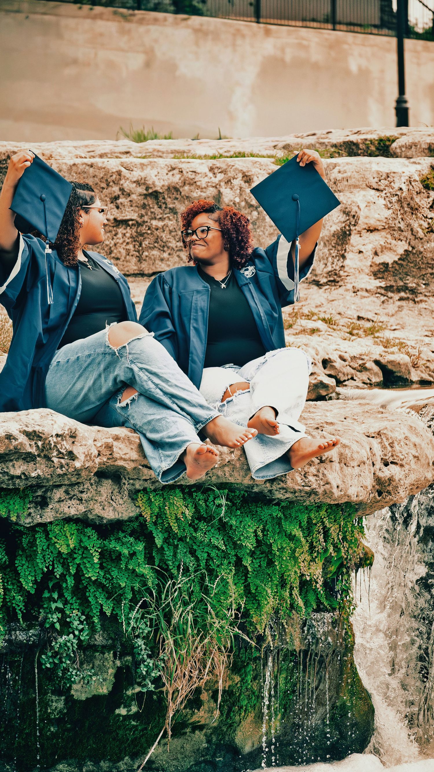 Two female graduates in blue robes sit on rocks holding caps near a mossy waterfall for senior photos.