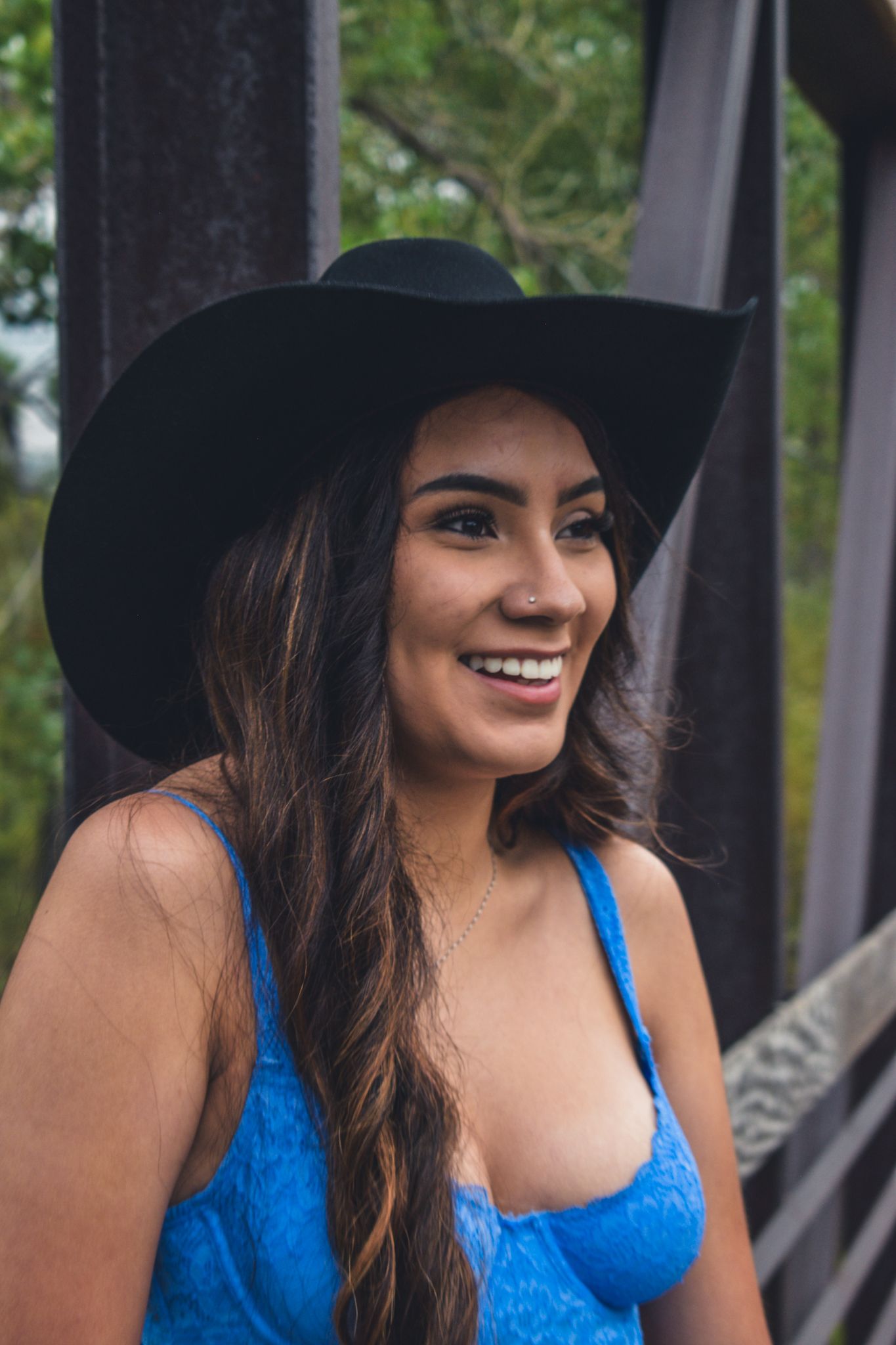 Smiling young woman in black cowboy hat and blue lace top poses on a rustic outdoor bridge for senior photos.