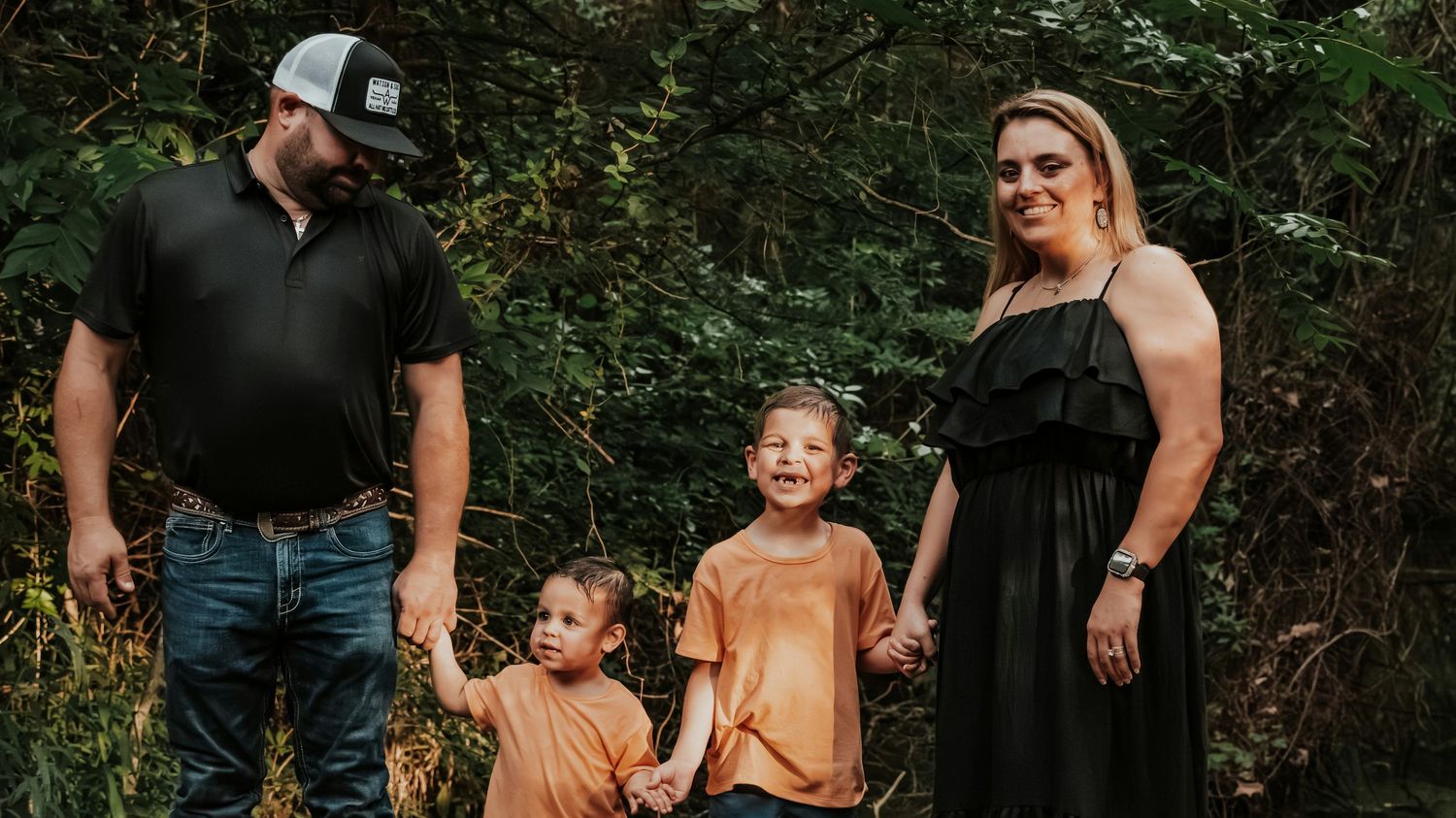 Family of four holding hands with two young boys in orange shirts walking together in a wooded area.