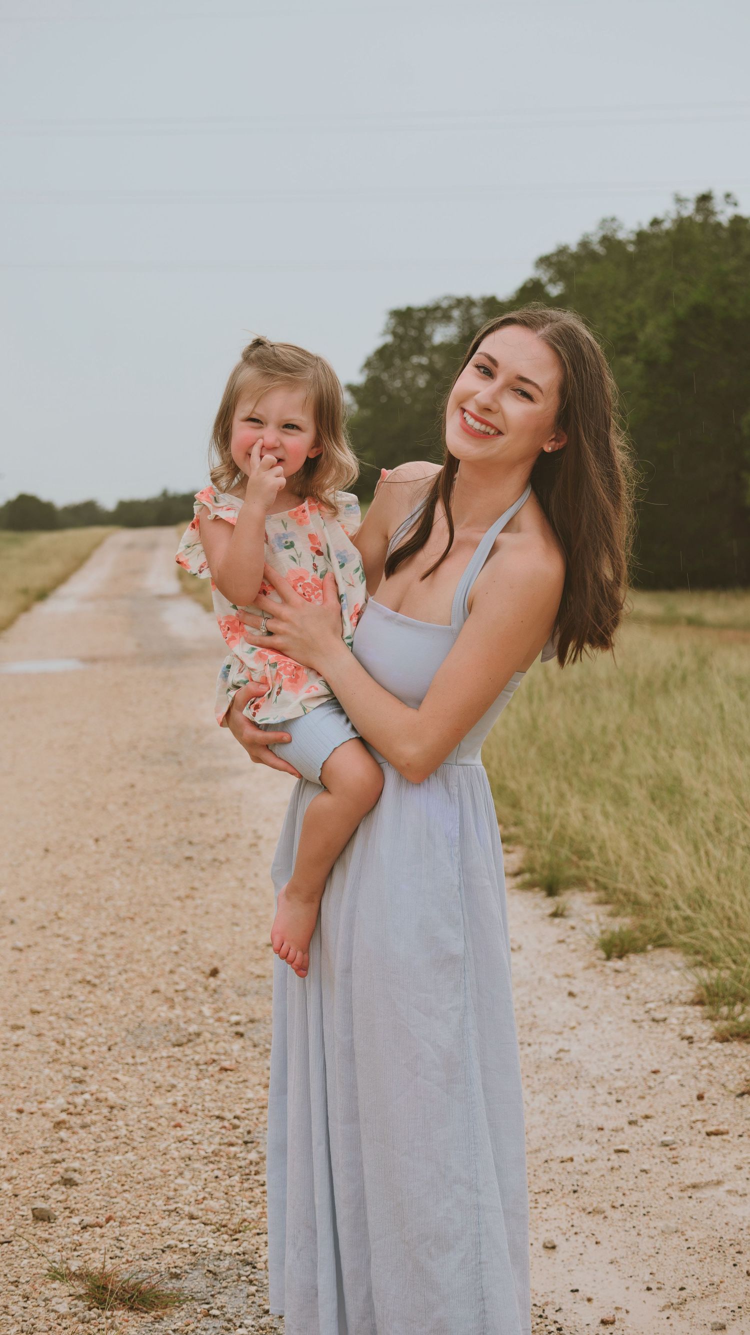 Beautiful young mother in gray dress smiles holding her toddler daughter outdoors.