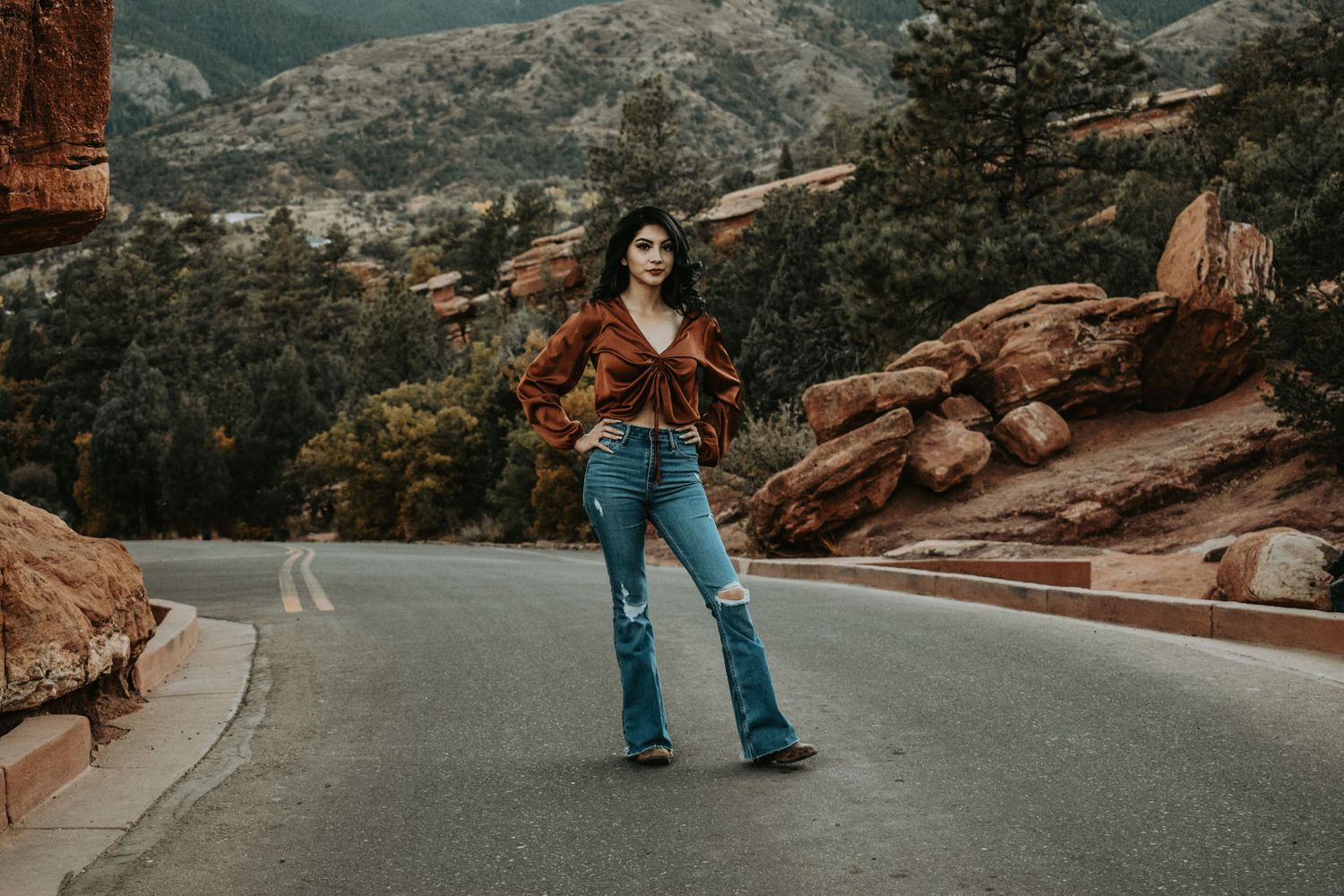 Young woman in rust blouse and flared jeans poses on mountain road surrounded by red rocks for senior photos.