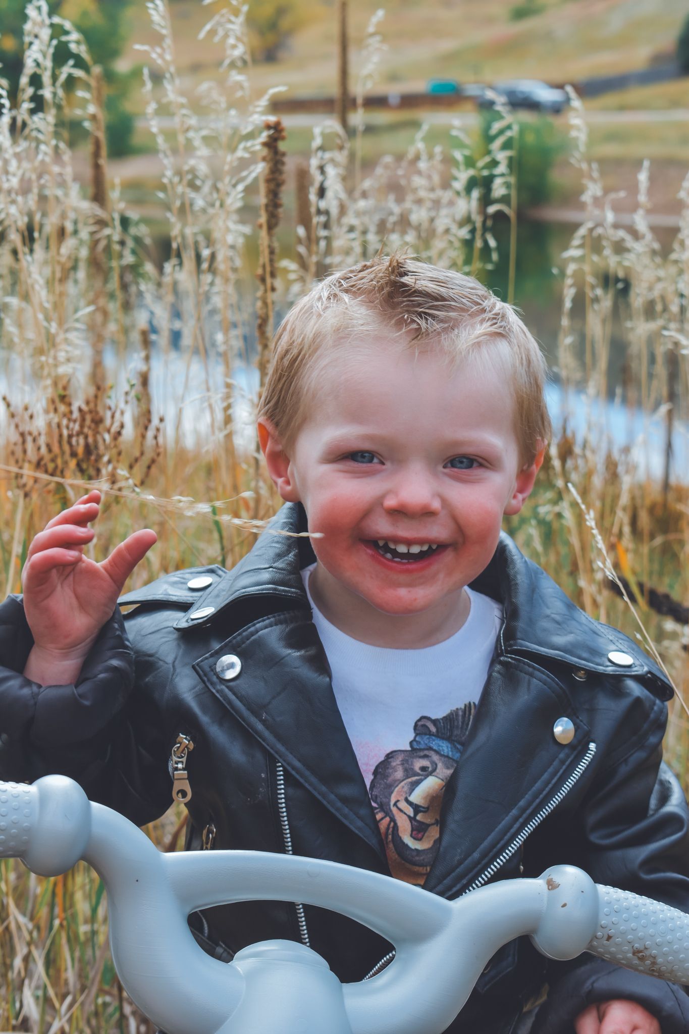 Joyful toddler in black leather jacket laughing and gesturing near toy bike in grassy field.