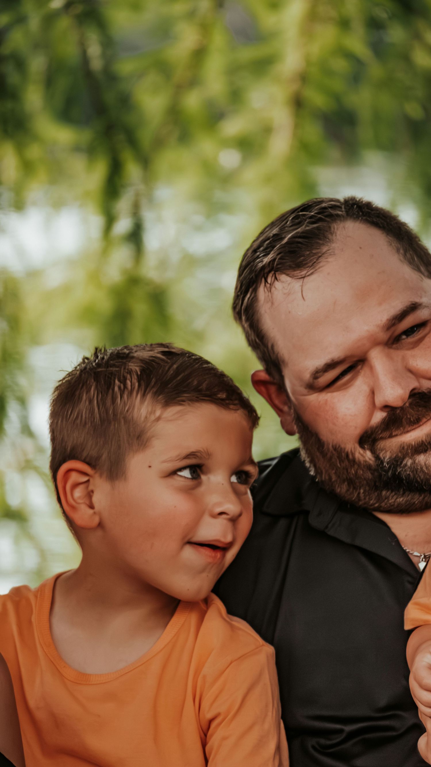 A bearded man in a black shirt smiles down at a young boy in an orange shirt outdoors near green trees.