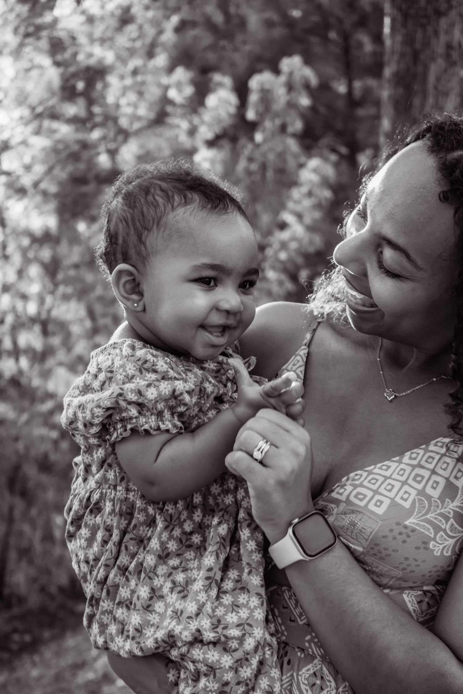 Black and white photo of a smiling mother holding her happy baby girl outdoors near blooming trees.