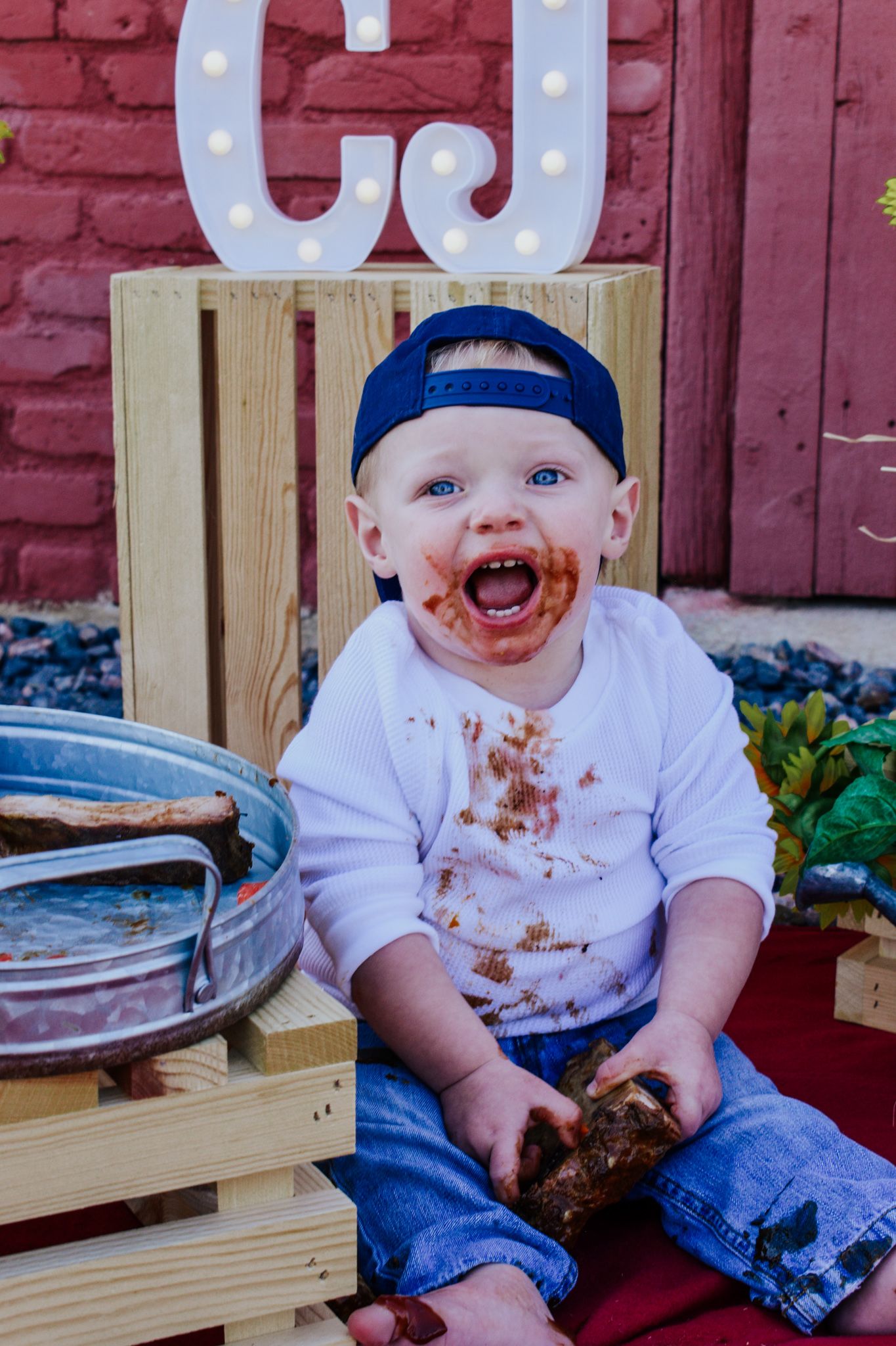 Laughing baby boy with chocolate cake smeared on face sits on wooden crate at birthday party. Family Photographer in Pflugerville, Tx