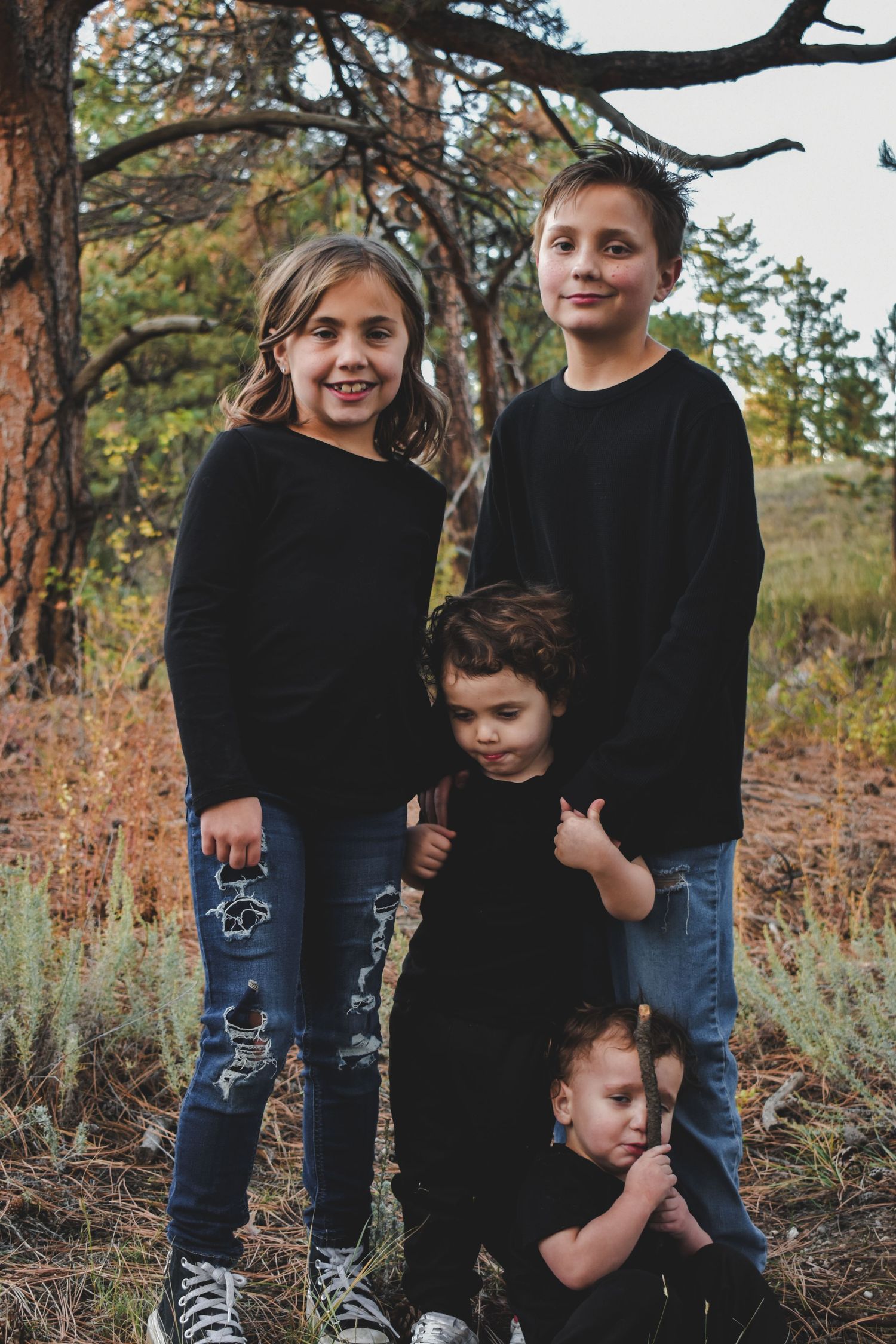 Four children in matching black tops and jeans pose together outdoors among autumn trees. Family Photographer in Pflugerville, Tx