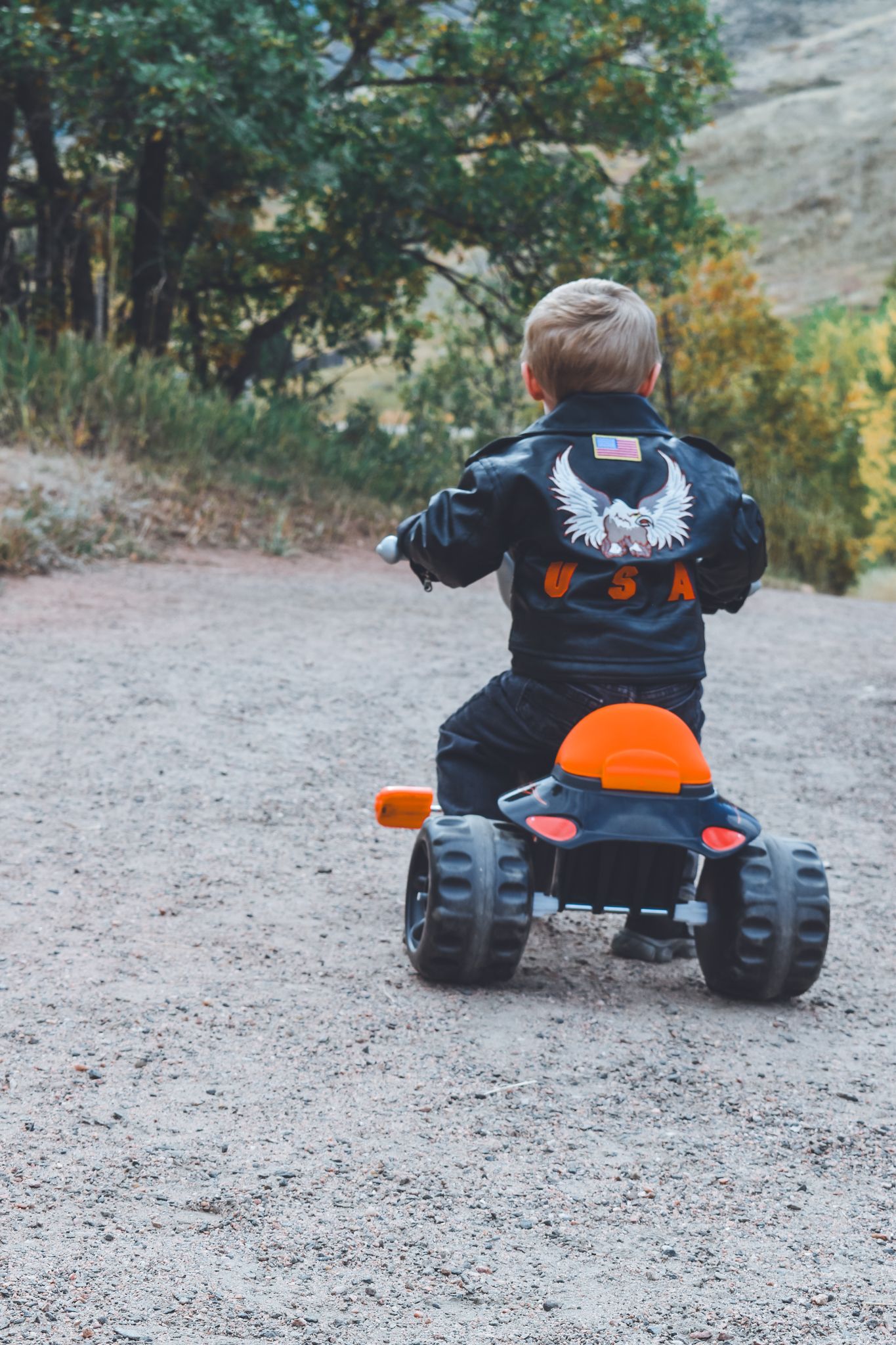 Young boy in USA leather jacket riding toy motorcycle on gravel path outdoors. Family Photographer in Pflugerville, Tx
