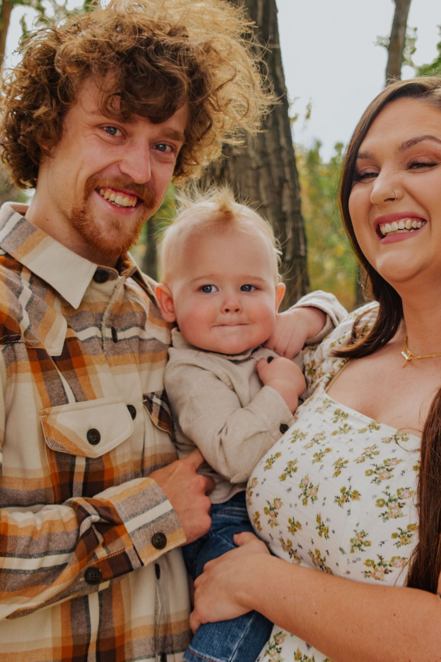 Smiling young family with curly-haired father, dark-haired mother, and blonde baby outdoors.