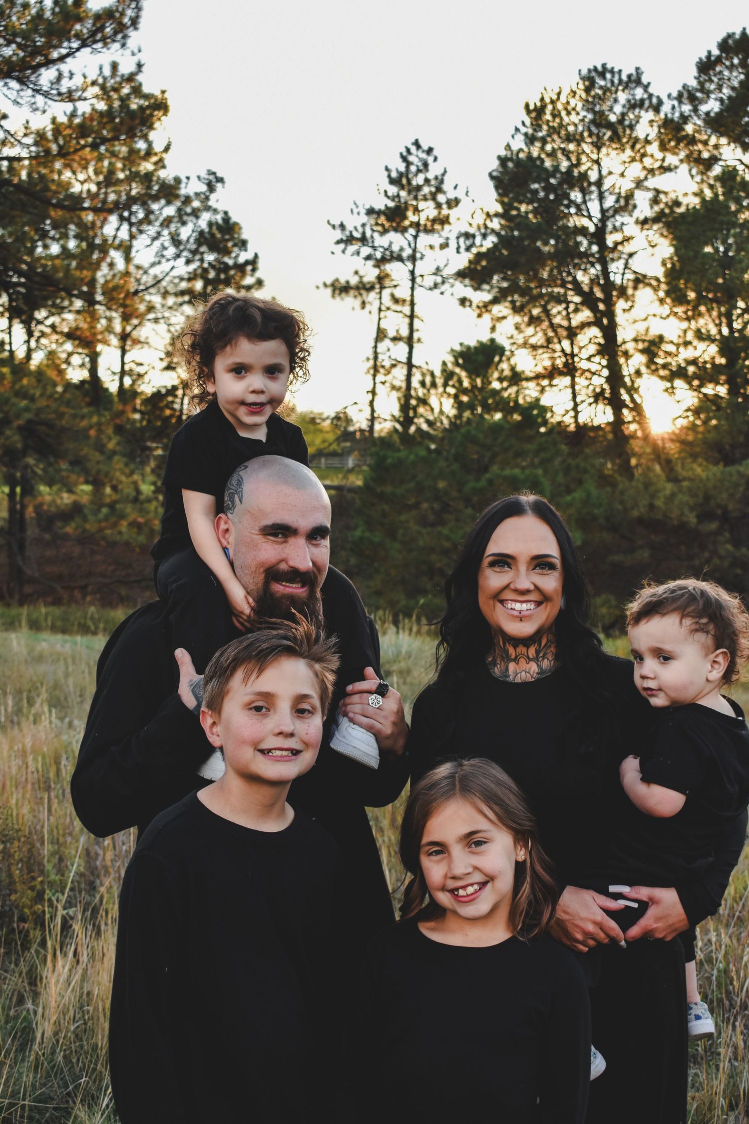 A family of six poses outdoors at sunset in matching black outfits, surrounded by tall pine trees in a grassy field.