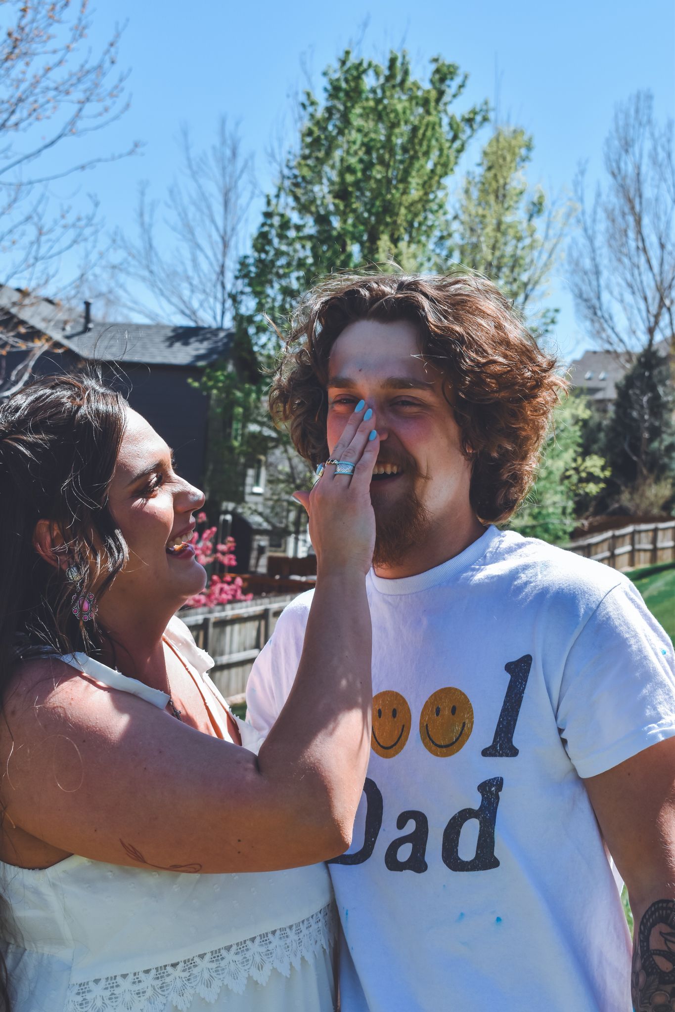 A laughing woman shows off her engagement ring to a smiling man wearing a Cool Dad t-shirt outdoors.