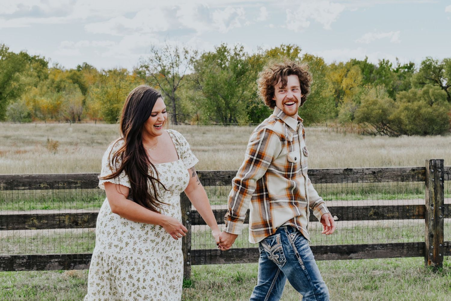 Laughing couple in casual country attire holding hands while walking along a rustic farm fence.
