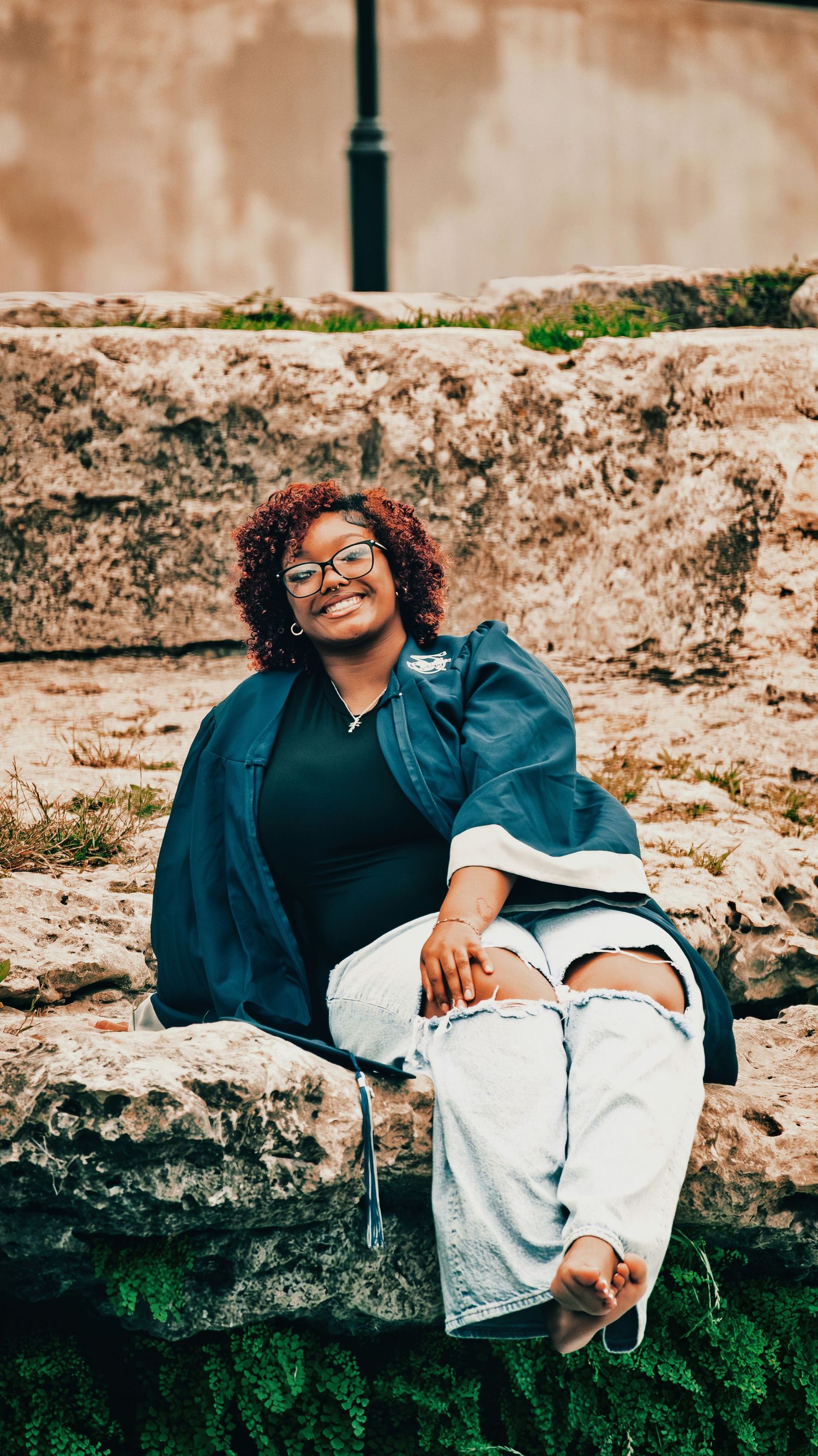 Young woman with curly hair and glasses smiling while sitting on a stone wall outdoors for senior photos.