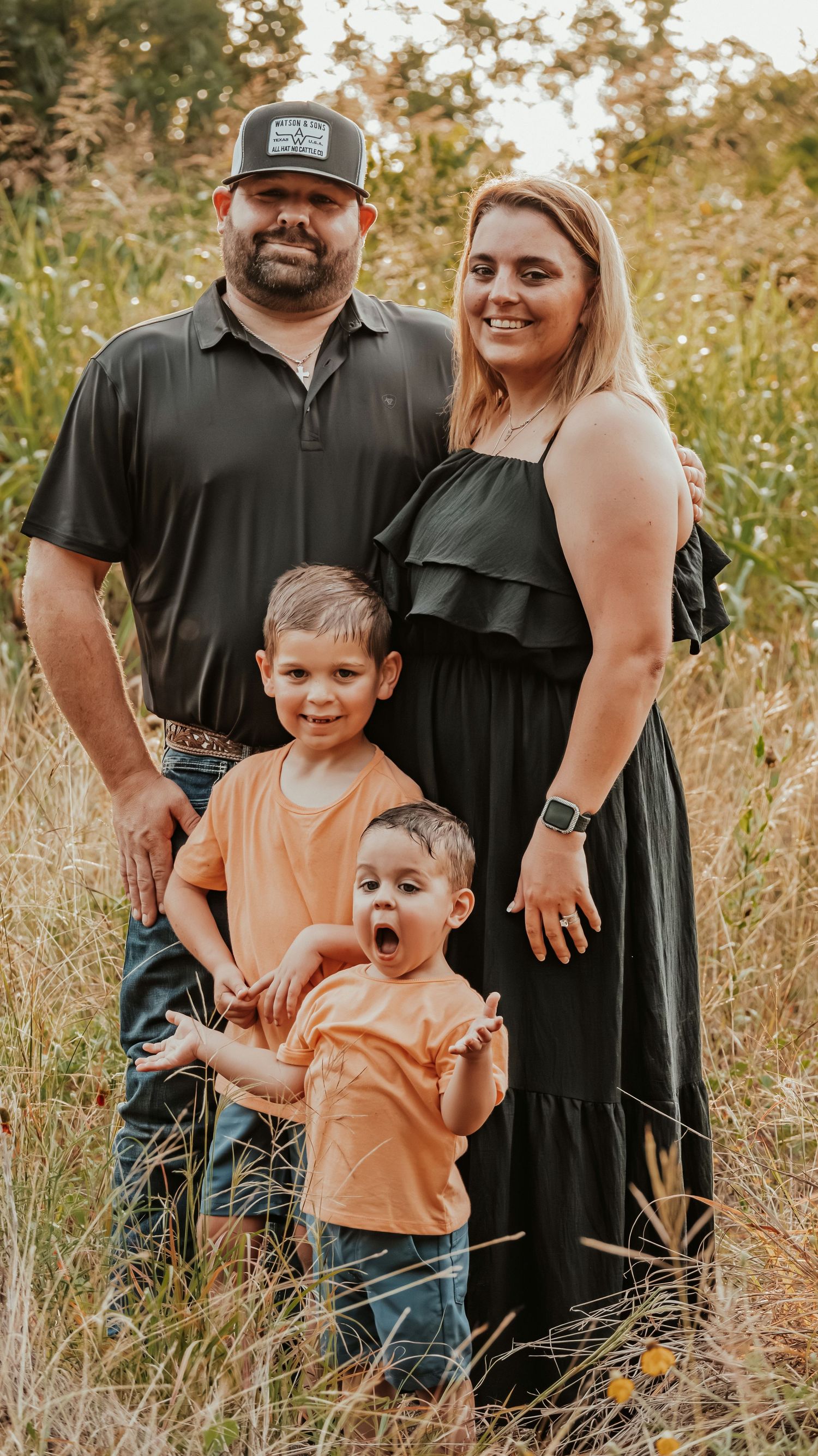 Family of four poses in golden field, parents in black outfits with two young boys in matching orange shirts.
