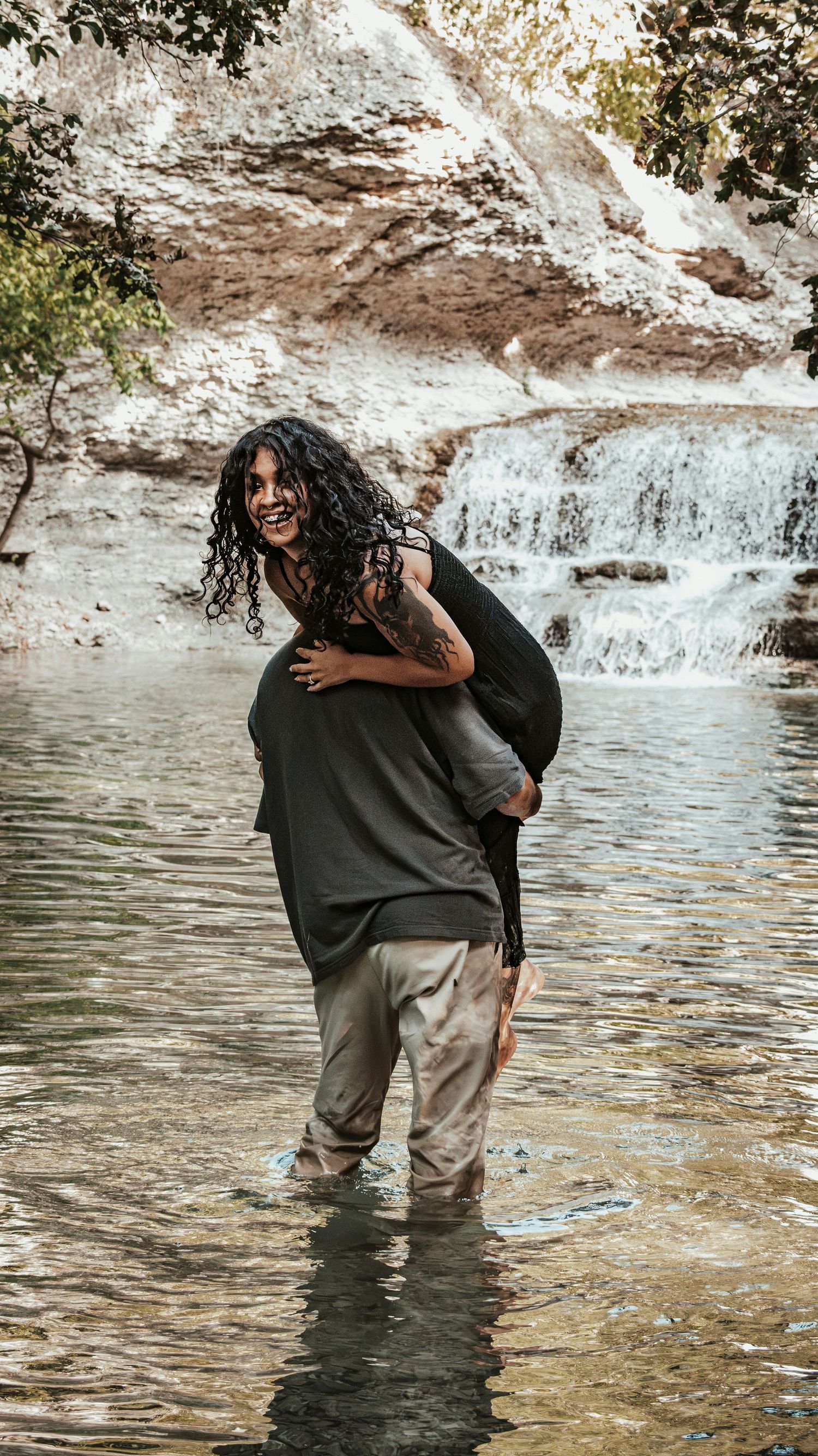 A laughing couple what a woman getting a piggyback ride in a shallow river with a waterfall for a couples session.