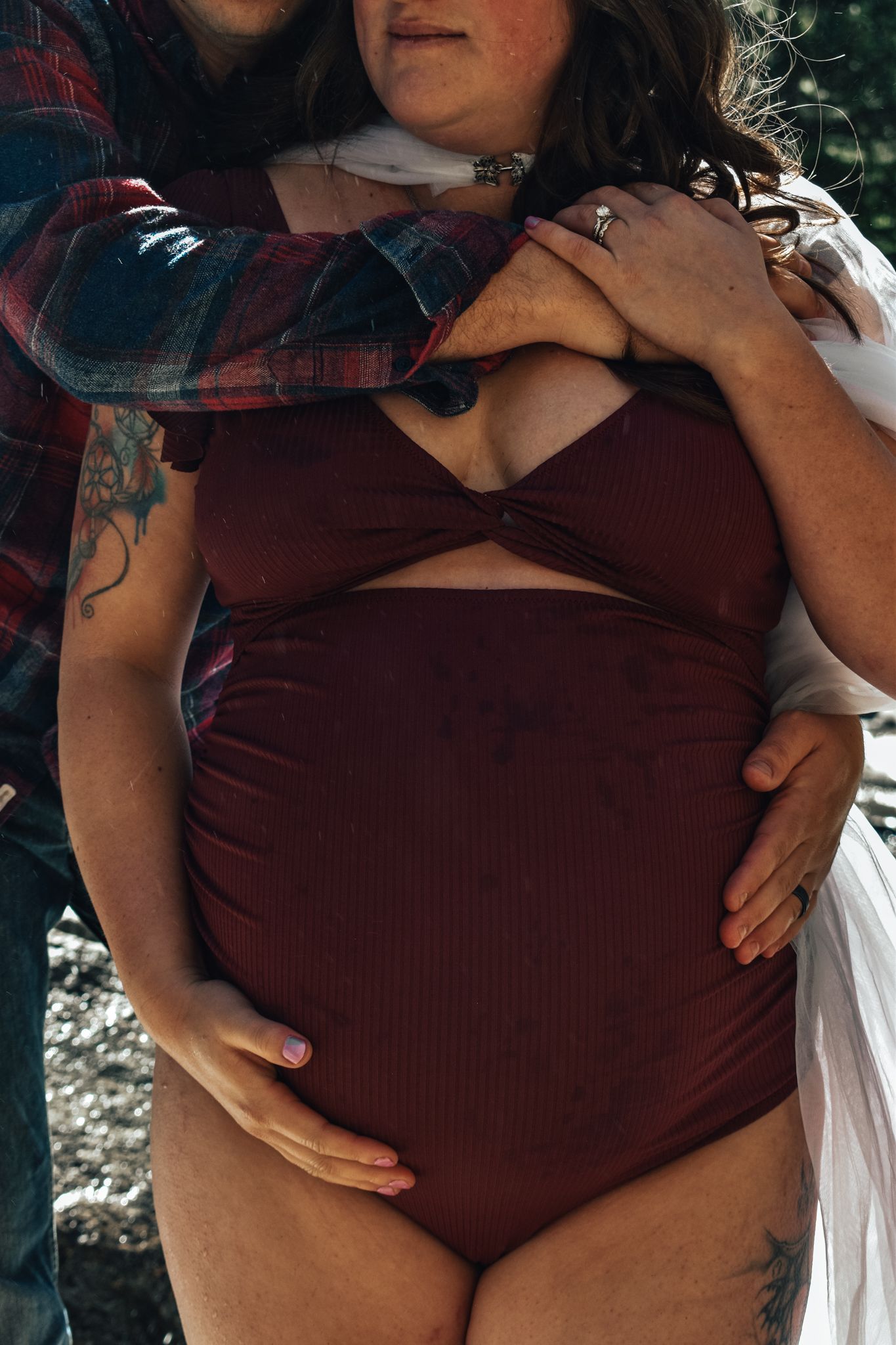 Pregnant woman in burgundy two-piece outfit being embraced from behind by partner outdoors. Maternity Photographer in Pflugerville, Tx