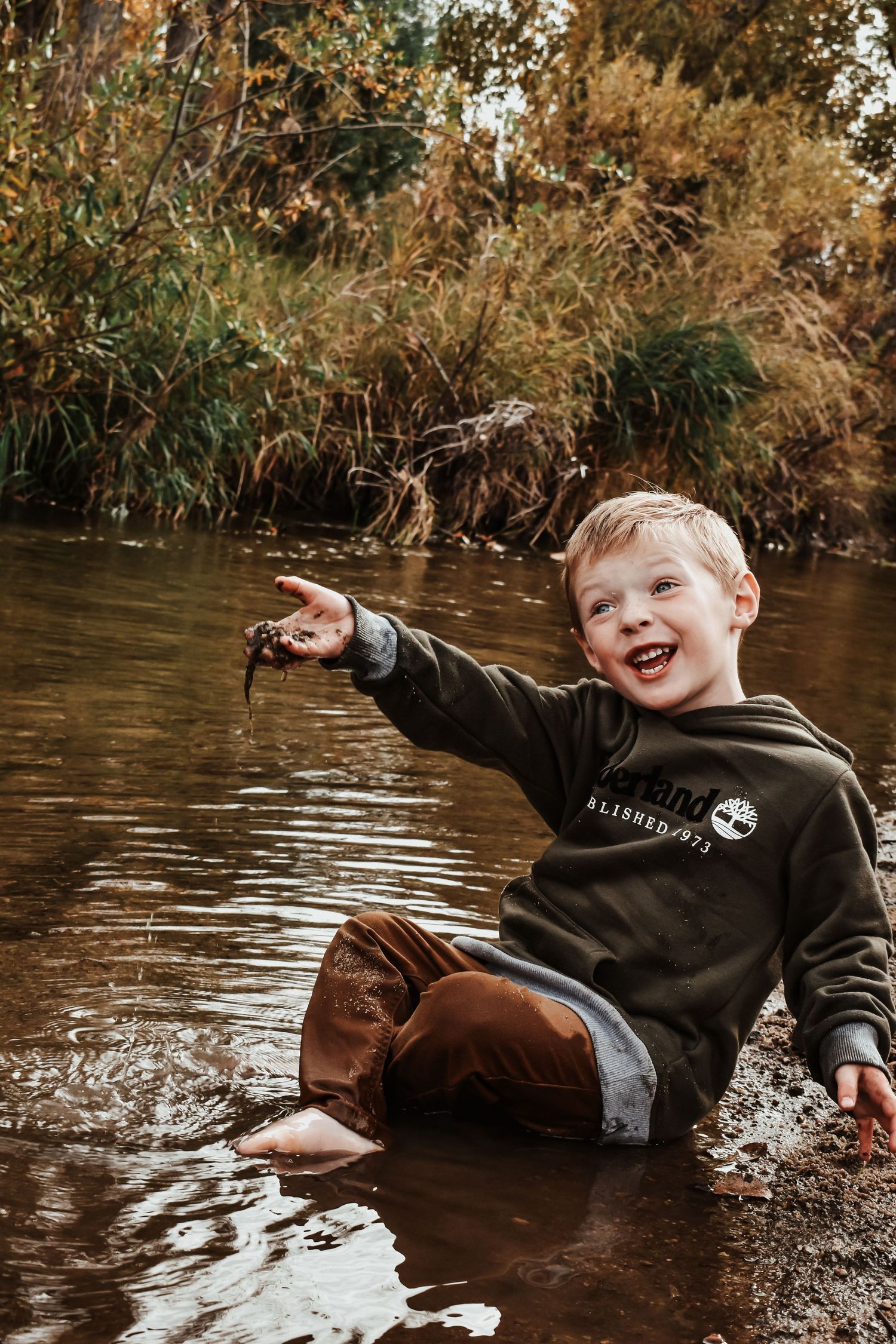 Laughing boy in Timberland hoodie sits in shallow creek water holding small creature outdoors. Family Photographer in Pflugerville, Tx
