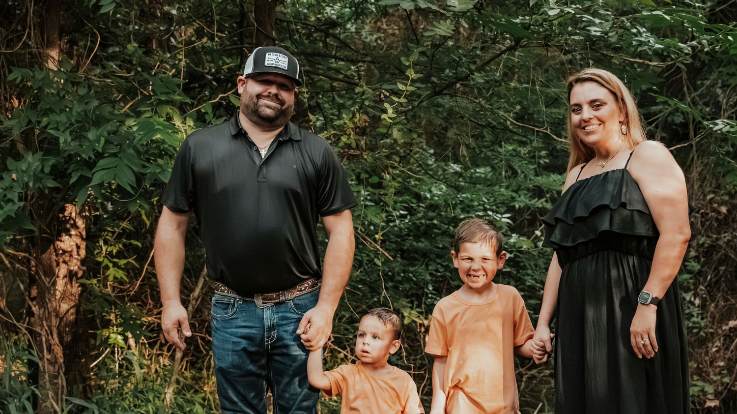 Family of four posing outdoors in nature, parents holding hands with two young boys wearing orange shirts.