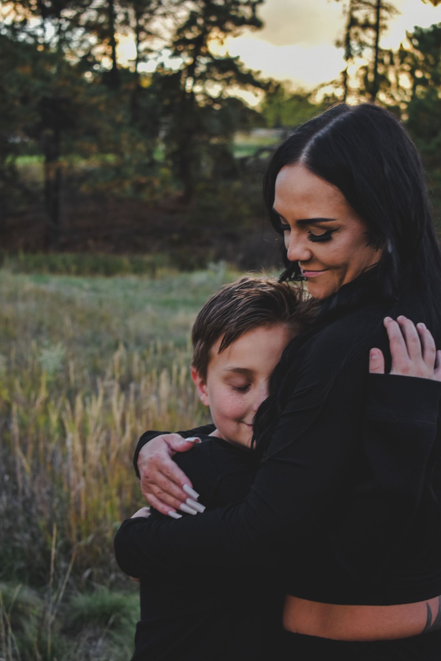Woman in black outfit warmly embracing a young boy in an outdoor field at sunset. Family Photographer in Pflugerville, Tx