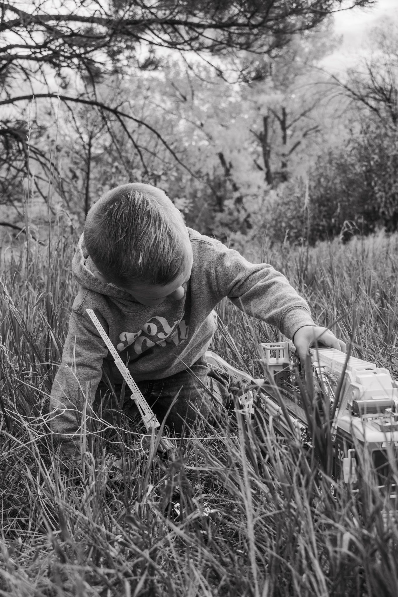 Black and white photo of young boy leaning over toy fire truck in tall grass surrounded by autumn trees.