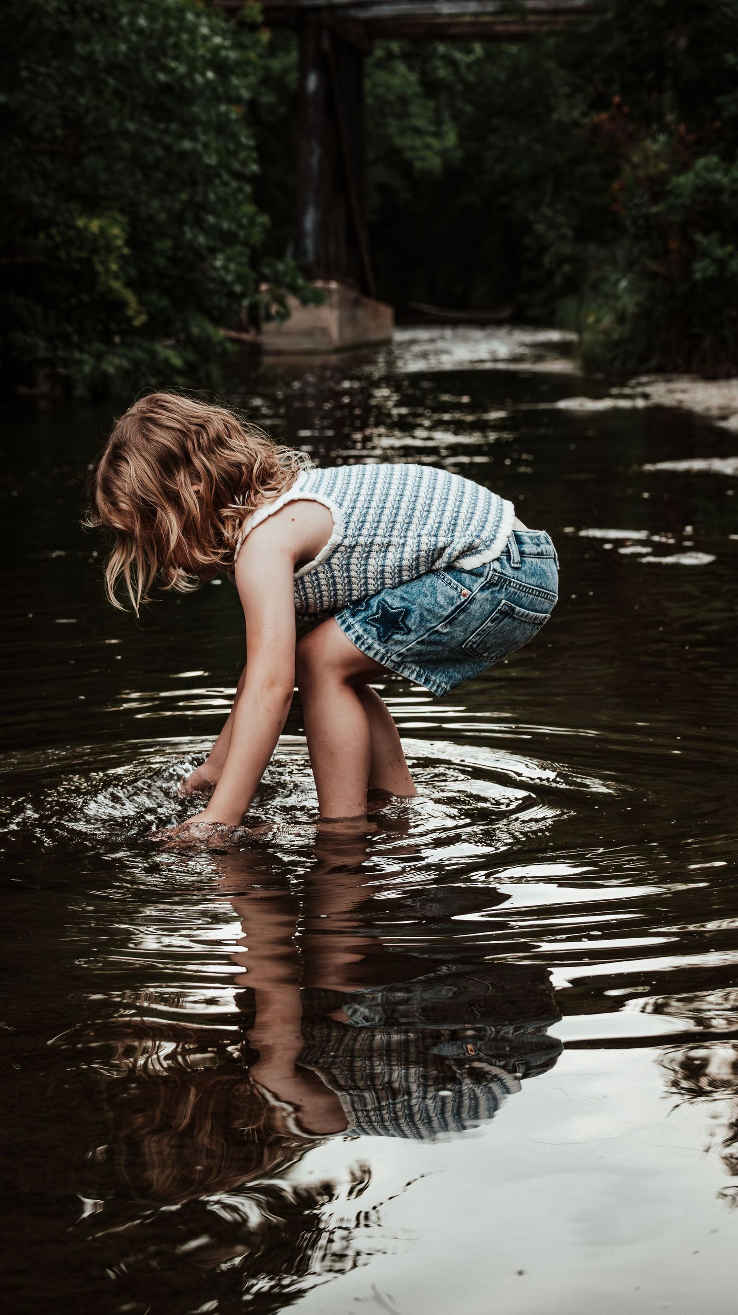 A young girl in a knit top and denim shorts bends down to touch the water in a creek.